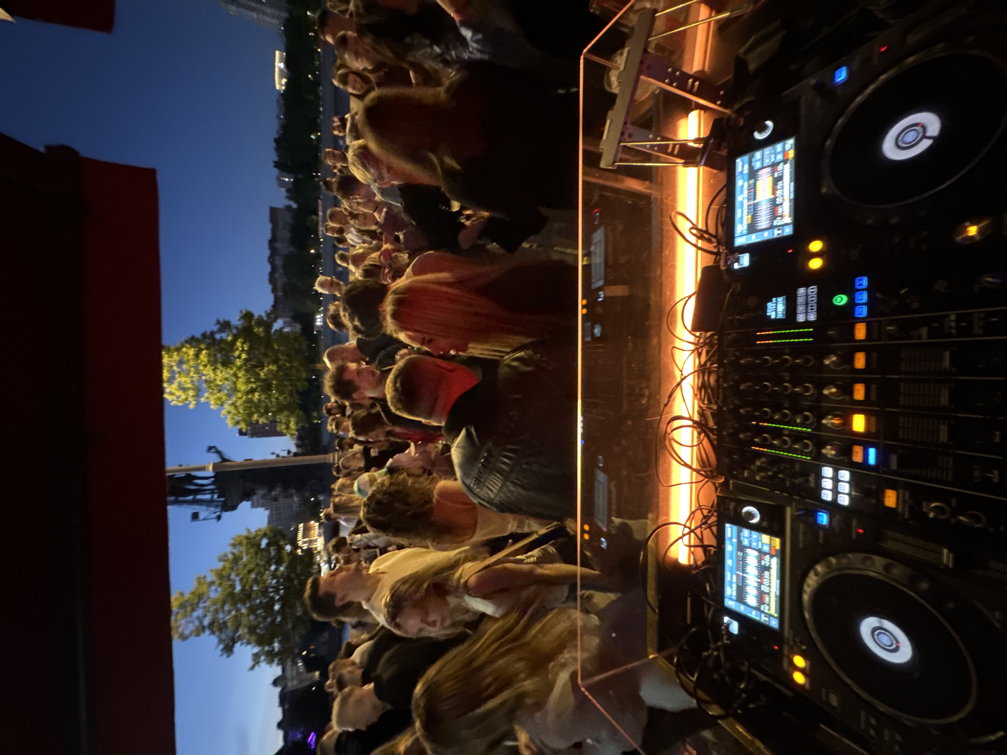 This image presents a detailed view of an outdoor evening social event along a riverbank in Moscow, Russia, viewed from behind a DJ console.
In the foreground, a professional DJ setup is visible, featuring two Pioneer CDJ-3000 multi-players and a Pioneer DJM series mixer, illuminated by an amber LED strip light running along the back of the console. Cables are haphazardly arranged behind the equipment. The screens of the CDJs display audio waveforms and track information. A transparent barrier, likely plexiglass, reflects parts of the crowd and the amber lighting.
The midground is densely populated with a diverse crowd of individuals, predominantly young adults. They are standing, conversing, and some appear to be dancing. Attire ranges from casual to semi-formal. Visible individuals include a man in a beige shirt facing right, a woman in a white top with long blonde hair looking downwards, and a man in a black leather jacket with his back to the camera, standing next to a woman with long blonde hair. General interactions indicate a lively social gathering.
The background reveals a dark blue twilight sky above a wide body of water, identified as the Moskva River. On the far bank, several buildings with illuminated windows are visible, interspersed with trees. A prominent, tall bronze monument, recognized as the "Monument to Peter the Great," stands distinctly on the distant riverbank, confirming the location as Moscow, Russia. The perspective suggests the event is taking place on an embankment or a moored vessel on the Moskva River, facing towards the monument.