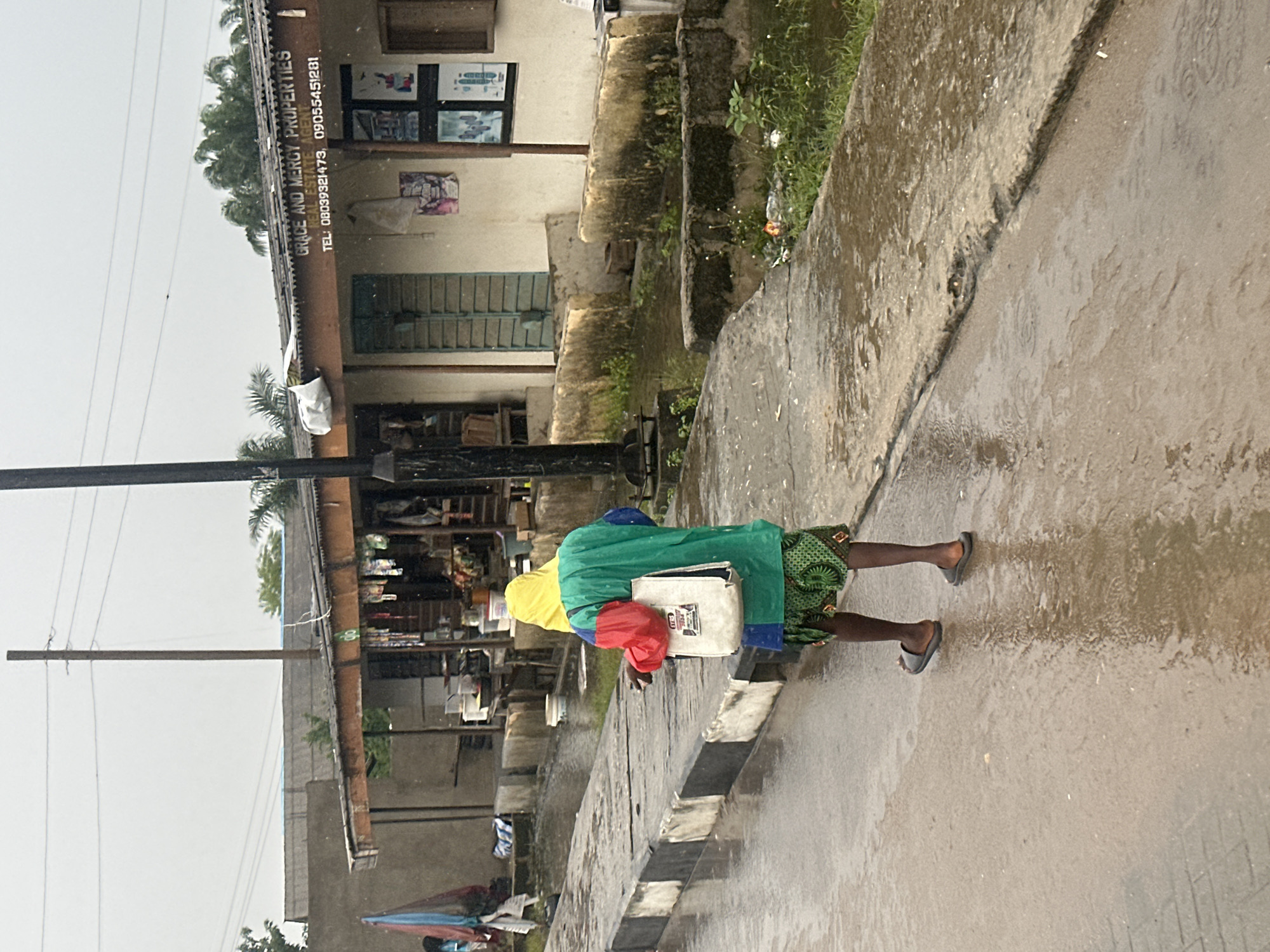 The media file depicts a person walking on a wet, paved surface, likely a street, in what appears to be a rainy or overcast day. The person is wearing a vibrant green and yellow jacket over a red shirt, and a patterned skirt. They are carrying a white bag. The setting is a roadside with a building visible to the left. The building has a corrugated metal roof and signs with text. One sign reads "GRACE AND MERCY PROPERTIES" and includes phone numbers "08039321473, 09055451281". The building's facade has windows and some posters or pictures displayed. To the right of the person, there is a grassy area with some debris and small plants. The ground is wet, with visible puddles and ripples, indicating recent or ongoing rain. The overall mood is somber due to the weather, but the bright clothing of the person adds a touch of color. The presence of overhead wires and a utility pole suggests an urban or semi-urban environment. The scene appears to be a street in Badagry, Nigeria.