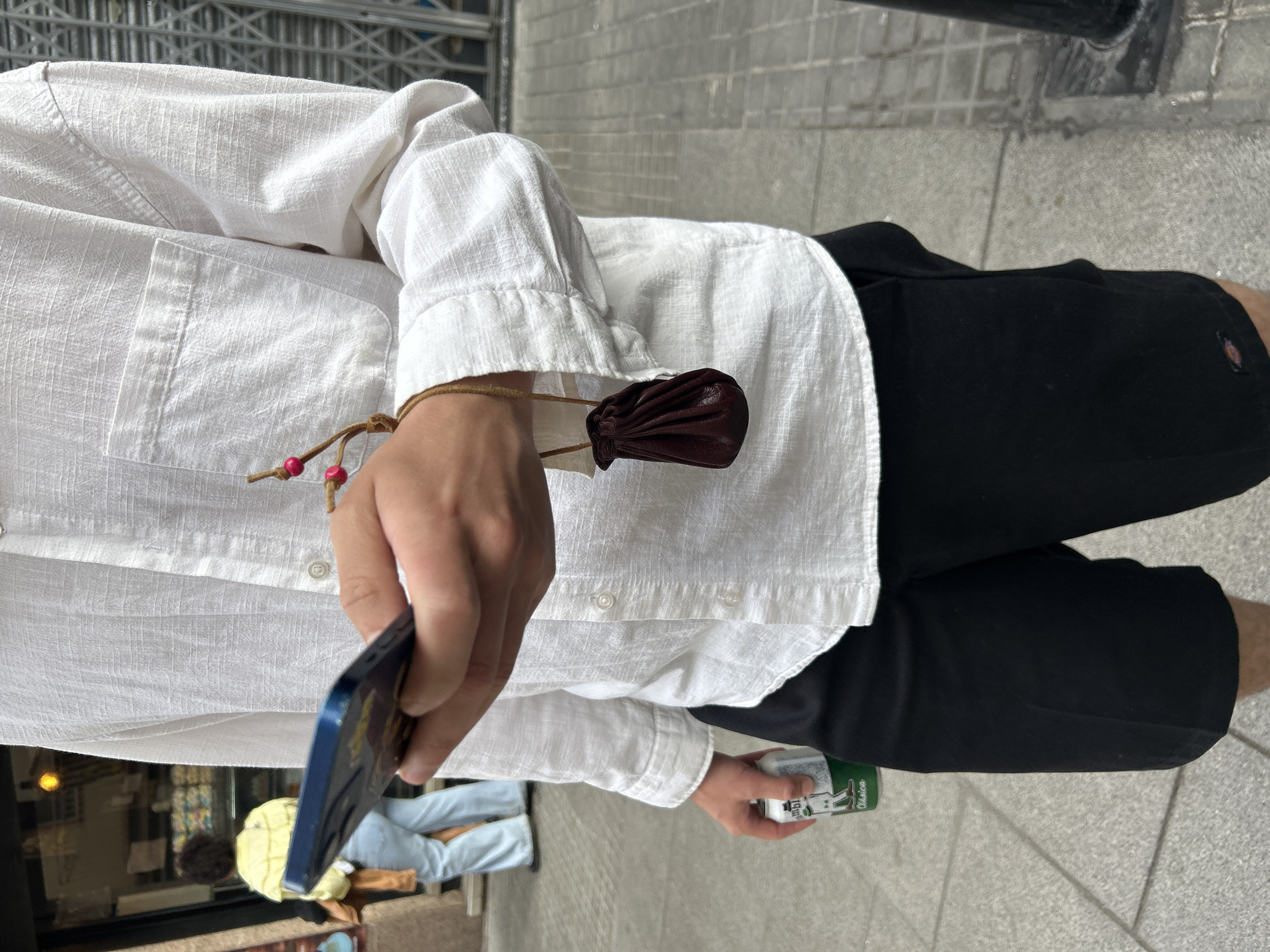A person wearing a white linen shirt and black shorts is standing on a tiled sidewalk. They are holding a dark-colored smartphone in one hand and a green and white can in the other. A small brown leather pouch is attached to the person's wrist with a drawstring. The location appears to be urban, with a building and people visible in the background, though partially obscured by the person's hand and phone.
