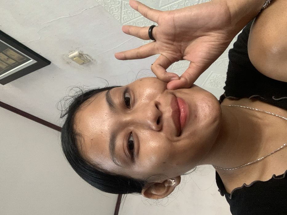 A woman is positioned at an unusual angle, seemingly taking a selfie from below. She has a dark ring on her finger and is pinching her cheek. She's wearing a black shirt with a scalloped neckline and a delicate silver necklace. The background includes a textured white ceiling, a framed picture hanging on the wall, and a light fixture.