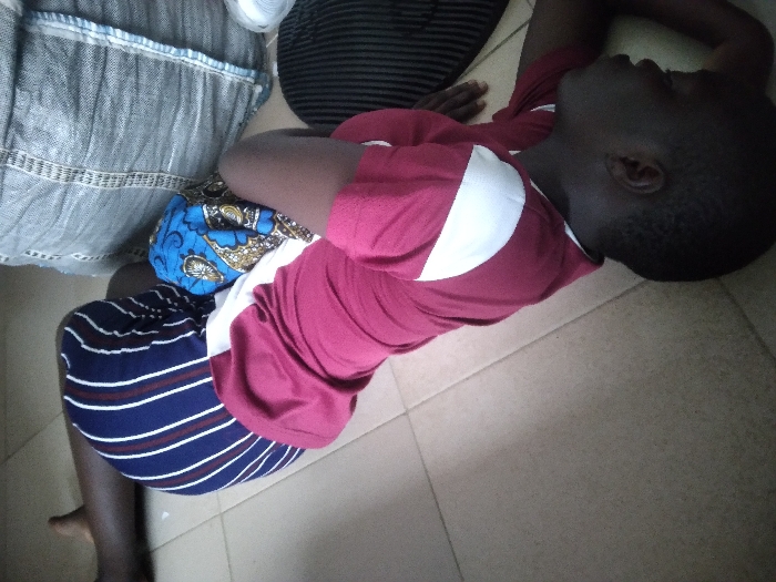 The image depicts an indoor scene in Agege, Nigeria, on a light-colored tiled floor. A dark-skinned individual lies supine with their head positioned towards the bottom right of the frame. This individual wears a maroon short-sleeved t-shirt featuring a white diagonal patch on the right shoulder/side. Their eyes are open, gazing upwards and to the right. Part of their lower body is covered by dark blue fabric with yellow and brown patterns, and one leg is bent at the knee. In the upper left corner, the bare lower leg and foot of a second dark-skinned individual are visible, alongside their upper leg covered by blue and white vertically striped trousers. A large grey woven sack rests on the floor near the upper body of the supine individual. To the right of the supine individual's head, a dark, possibly ribbed, cap or hat and a small roll of white material are positioned on the tiled surface. The floor is composed of light beige or cream square tiles with dark grout lines.