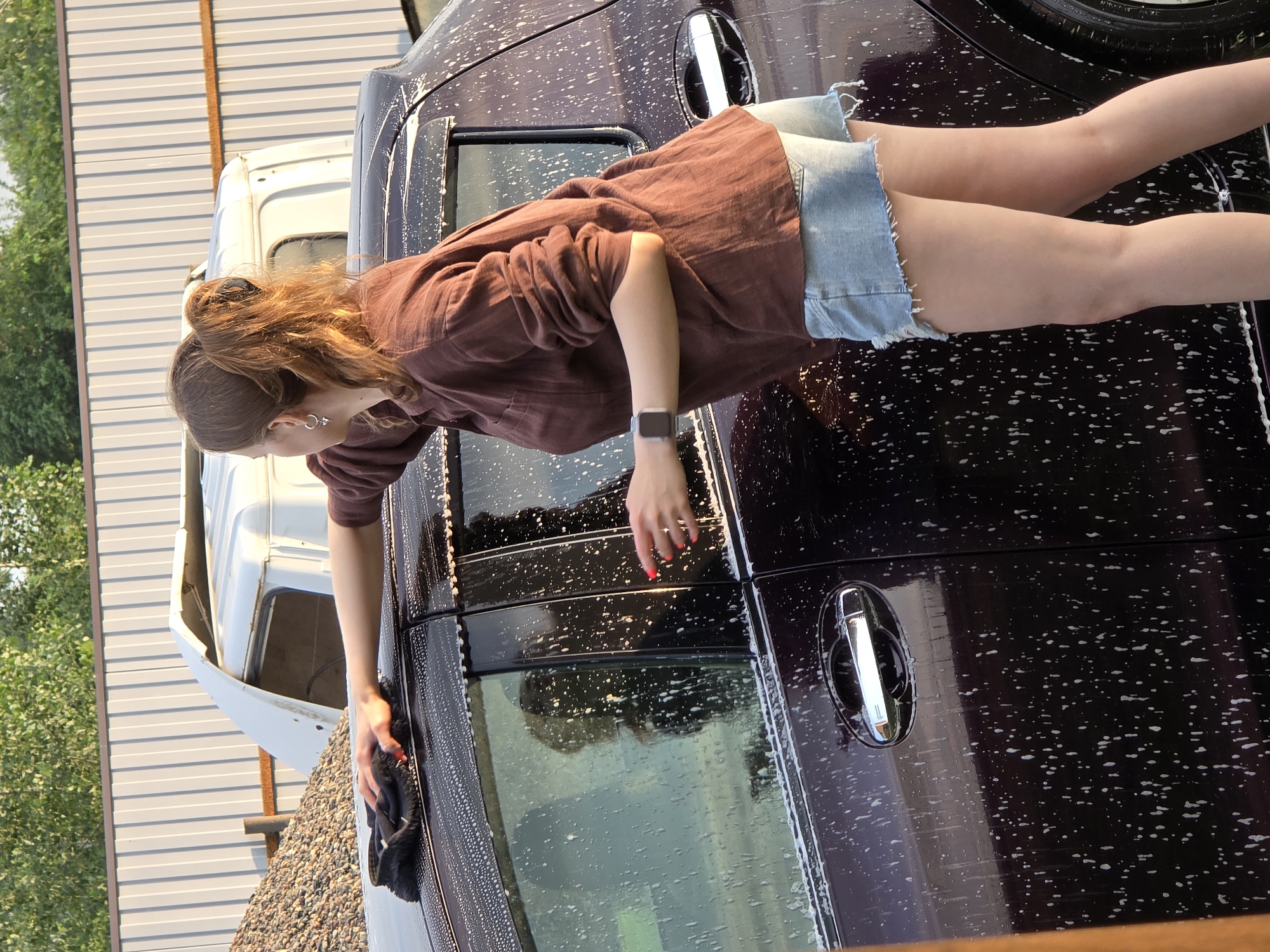 A young woman is washing a dark-colored car, which is covered in soap suds. She is wearing a brown linen shirt and denim shorts. Her hair is pulled back in a bun. She is holding a black wash mitt in her right hand and appears to be wiping down the car door. A watch is visible on her left wrist. The car is parked next to a white truck and a building with corrugated metal siding. The scene is outdoors, and the lighting suggests it is daytime, possibly late afternoon or early evening, with the sun casting long shadows. Green foliage is visible in the background. The overall impression is that of a casual, domestic activity. The location is identified as Ermolaevo, Russia. There is no visible text in the image.