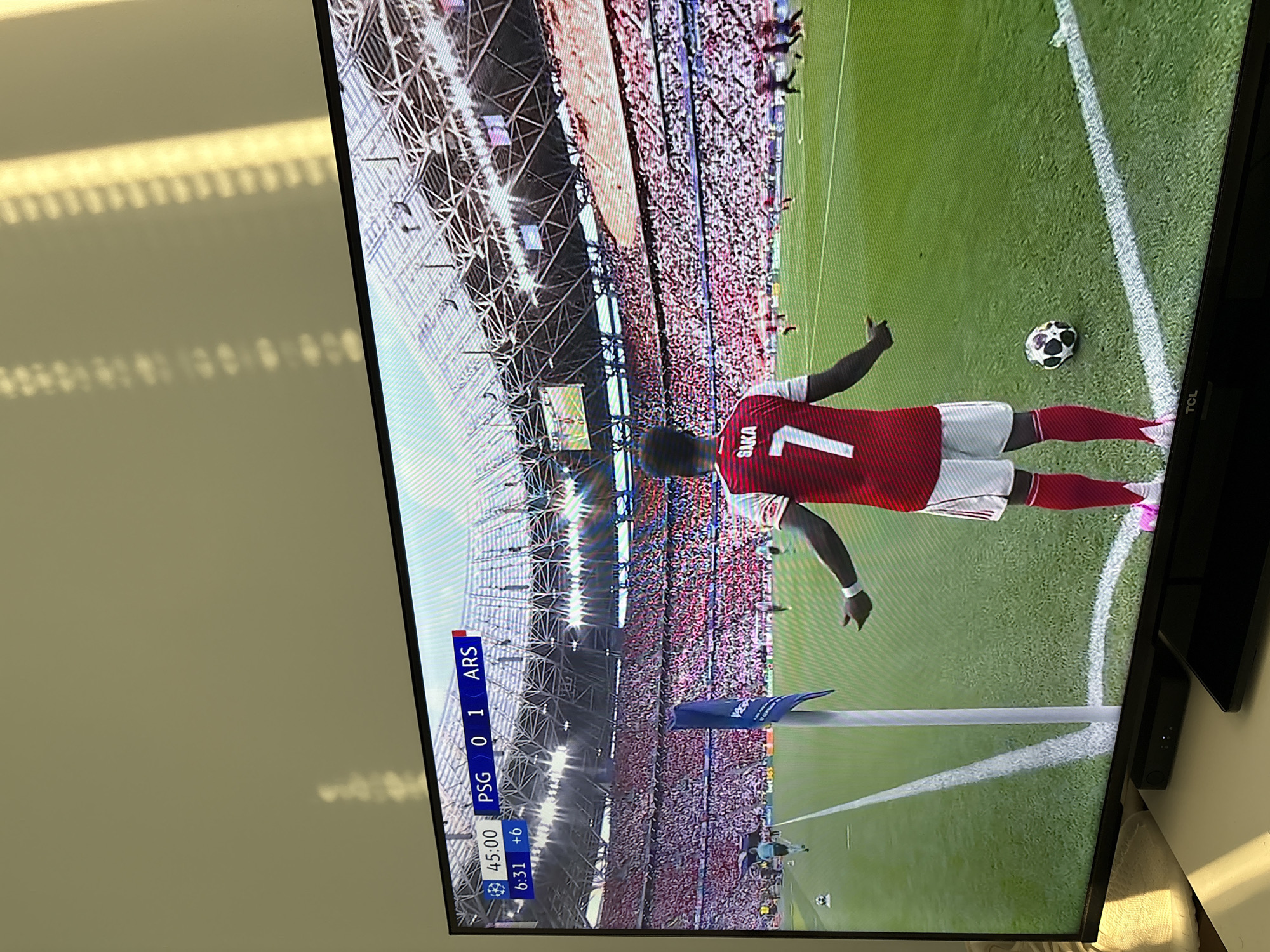 The image shows a television screen displaying a soccer video game. The score is PSG 0, ARS 1. The time is 45:00 +6. A player wearing an Arsenal jersey with the number 7 and "SAKA" on the back is standing near the corner flag. A soccer ball is visible nearby on the grass field. A large crowd of spectators fills the stadium. The game appears to be a Champions League match. The location is a soccer stadium in an unknown city.