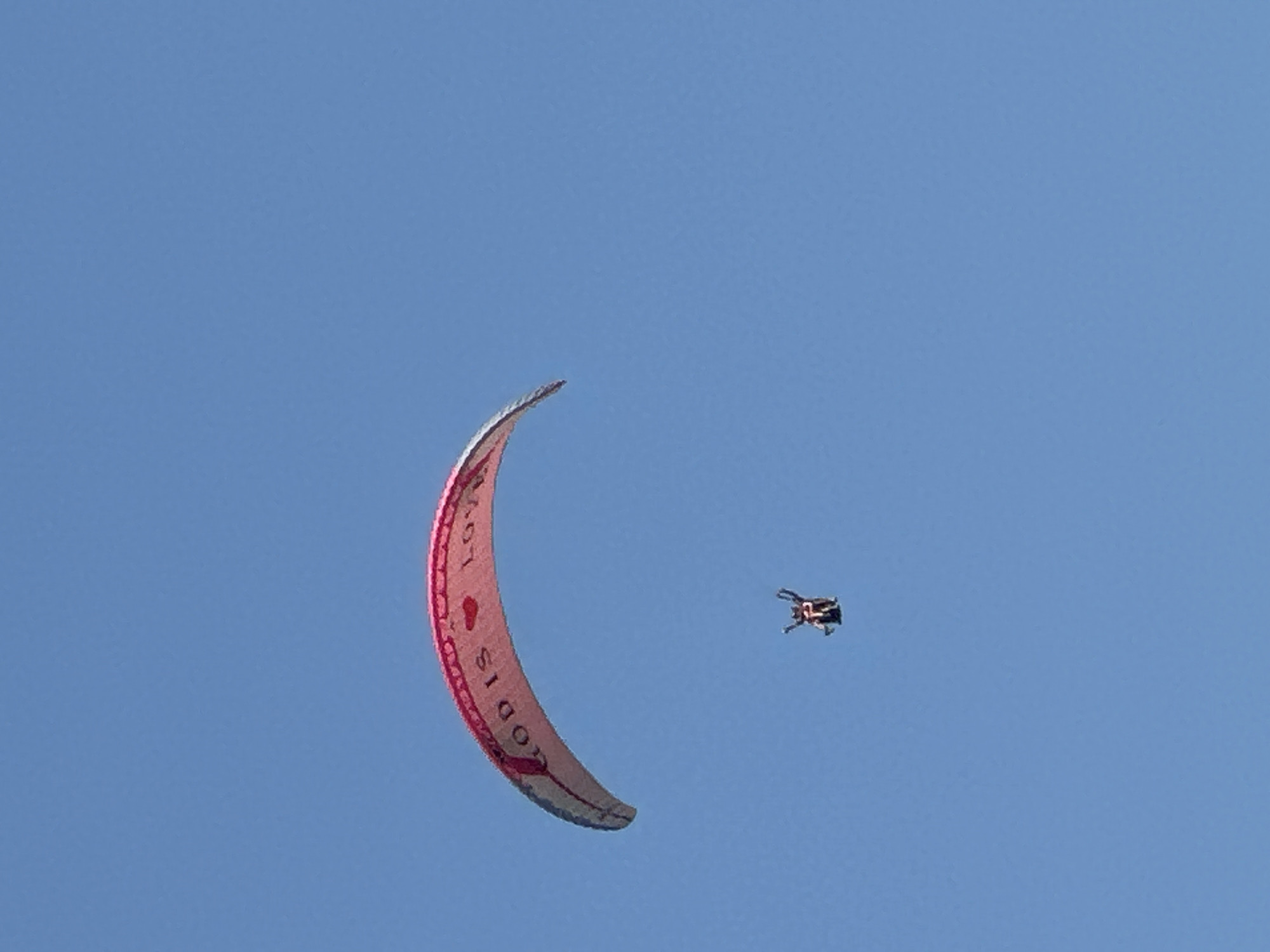A paraglider with the text "GOD IS LOVE" and a heart symbol printed on its canopy is soaring against a clear blue sky. Below the paraglider, a person is suspended in a harness, with their arms raised in a celebratory gesture. The scene is captured from a low angle, emphasizing the vastness of the sky and the height of the paraglider and pilot. No specific city or geographical markers are visible in the image to determine the location.