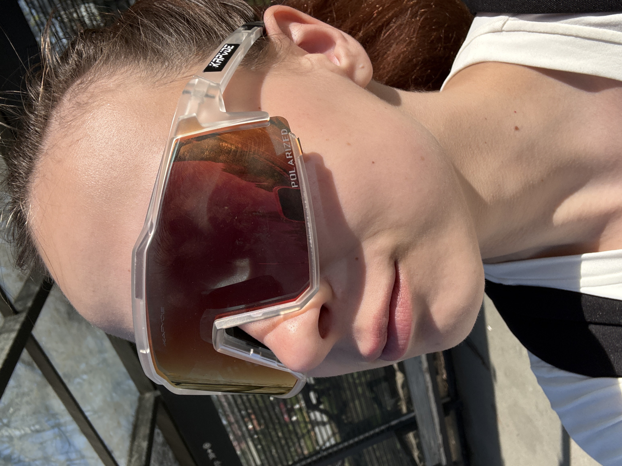 A person with light skin lies down outside, wearing sunglasses with a translucent frame and polarized lenses that reflect the surrounding environment. The sunglasses have the brand name "KRPDE" printed on the side. The person has brown hair pulled back. They are wearing a white shirt with a black stripe visible near the shoulder. The background appears to be an urban environment with metal structures and foliage. The location is identified as Perm, Russia, based on the reflection in the sunglasses lenses, which feature distinctive Perm architecture.