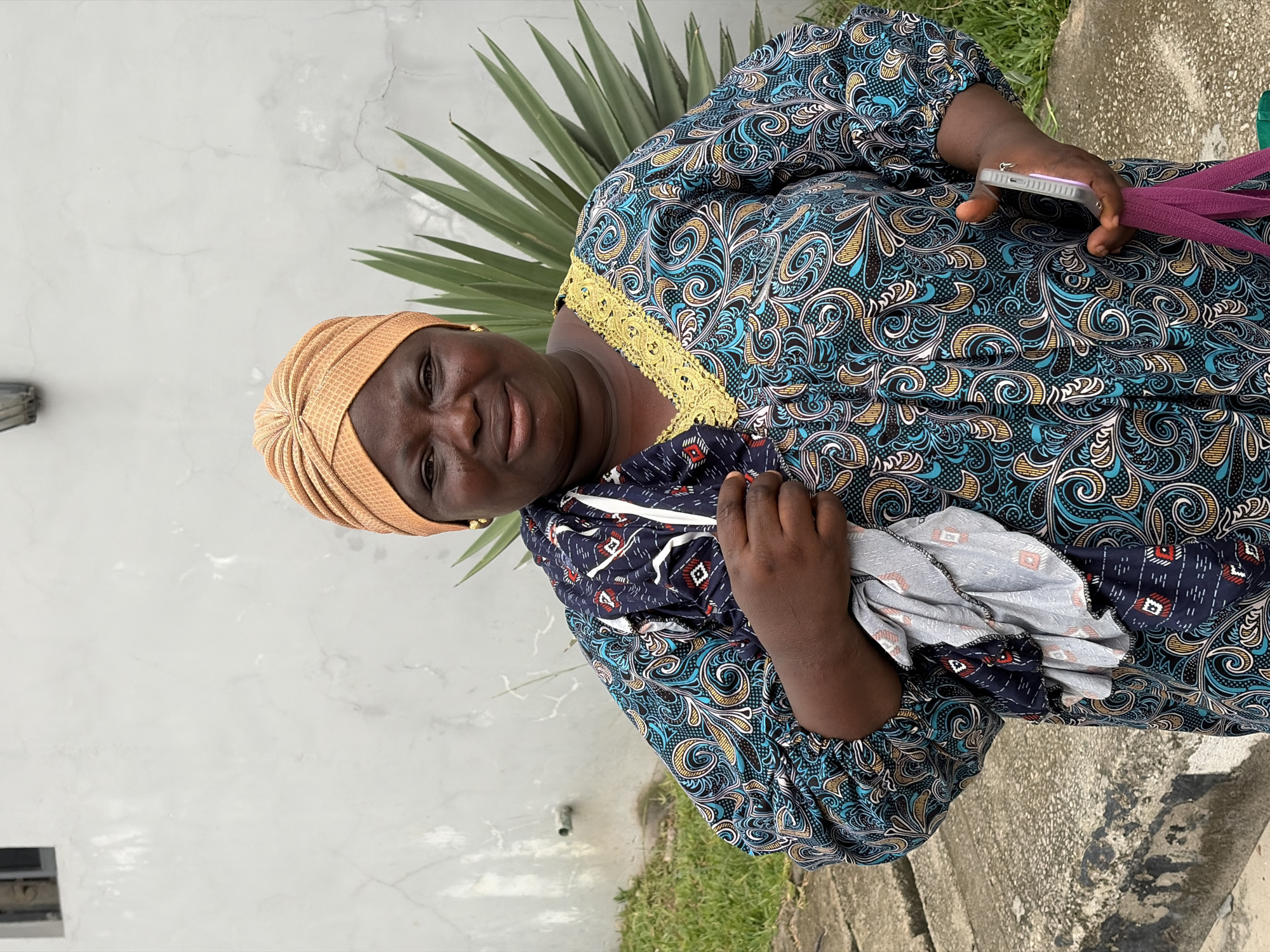 A woman smiles gently towards the camera in an outdoor setting in Igboefon, Nigeria. She is positioned against a light grey, somewhat cracked wall, with a tall, spiky-leaved tropical plant visible behind her.
The woman has a dark complexion and wears a light orange, pleated headwrap. Her attire includes a long-sleeved dress featuring an elaborate pattern of swirling motifs and paisley designs in dark blue, light blue, gold, and white on a dark background. The neckline of the dress is accentuated with a golden, embroidered trim.
In her right hand, she holds a light-colored smartphone and a purple strap, possibly from a bag. Her left hand holds a folded piece of fabric, which is dark blue with small red and white geometric patterns, resembling the fabric of another garment or accessory.
The scene is brightly lit, indicating daytime and clear weather. The woman's expression is warm and inviting. At the bottom of the frame, a concrete surface or pavement is partially visible. There is no discernible text within the image.