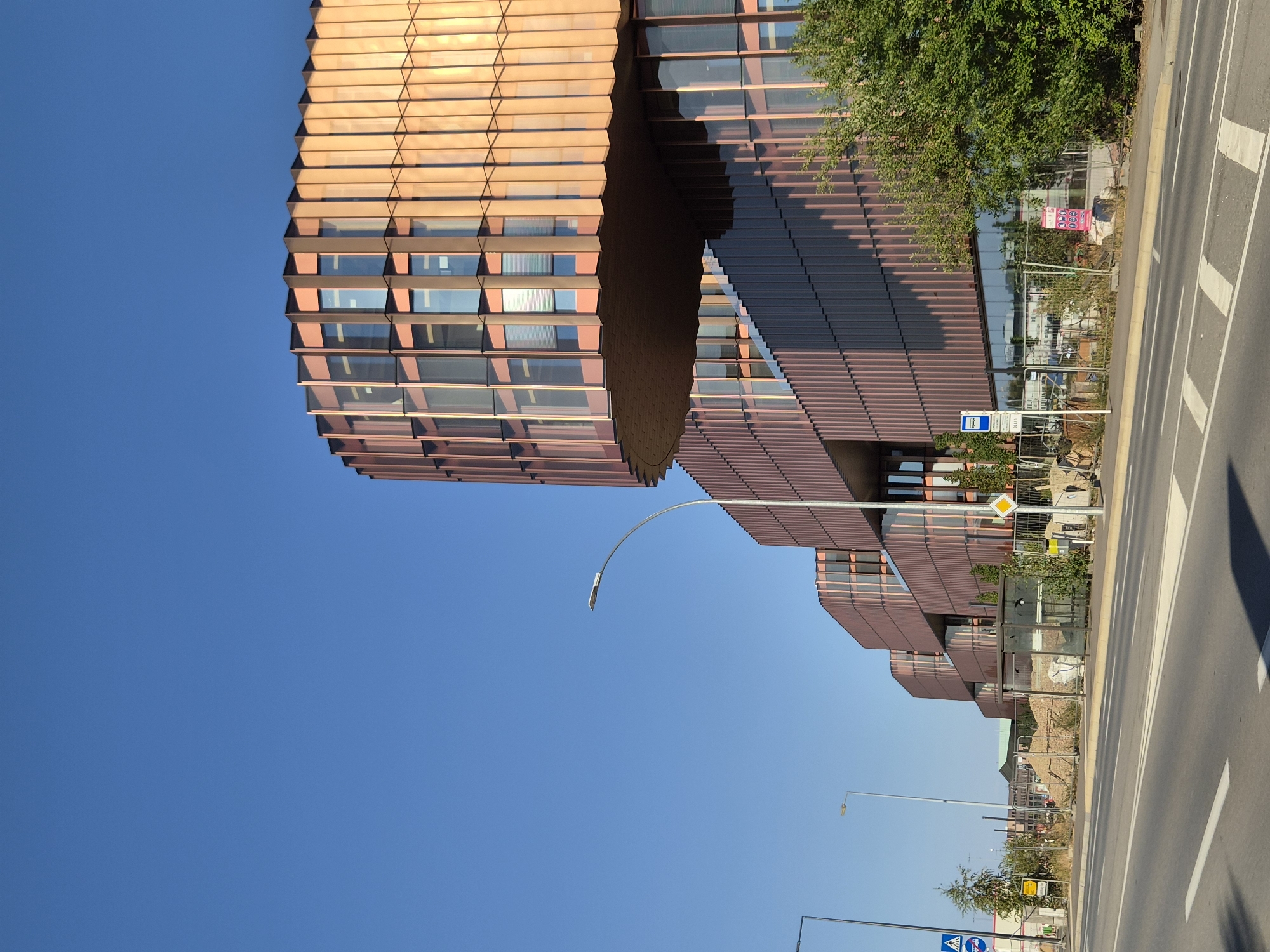 The image captures a modern, multi-story building with a distinctive curved facade made of reddish-brown vertical slats and glass panels. The building appears to be either under construction or recently completed, as evidenced by piles of dirt and construction fencing along the side. The structure is situated next to a paved road with white lane markings and a crosswalk. A bus stop shelter, a street light, and a few trees are visible in front of the building. The sky is clear and blue, suggesting a sunny day. The scene is located in Niederanven, Luxembourg.