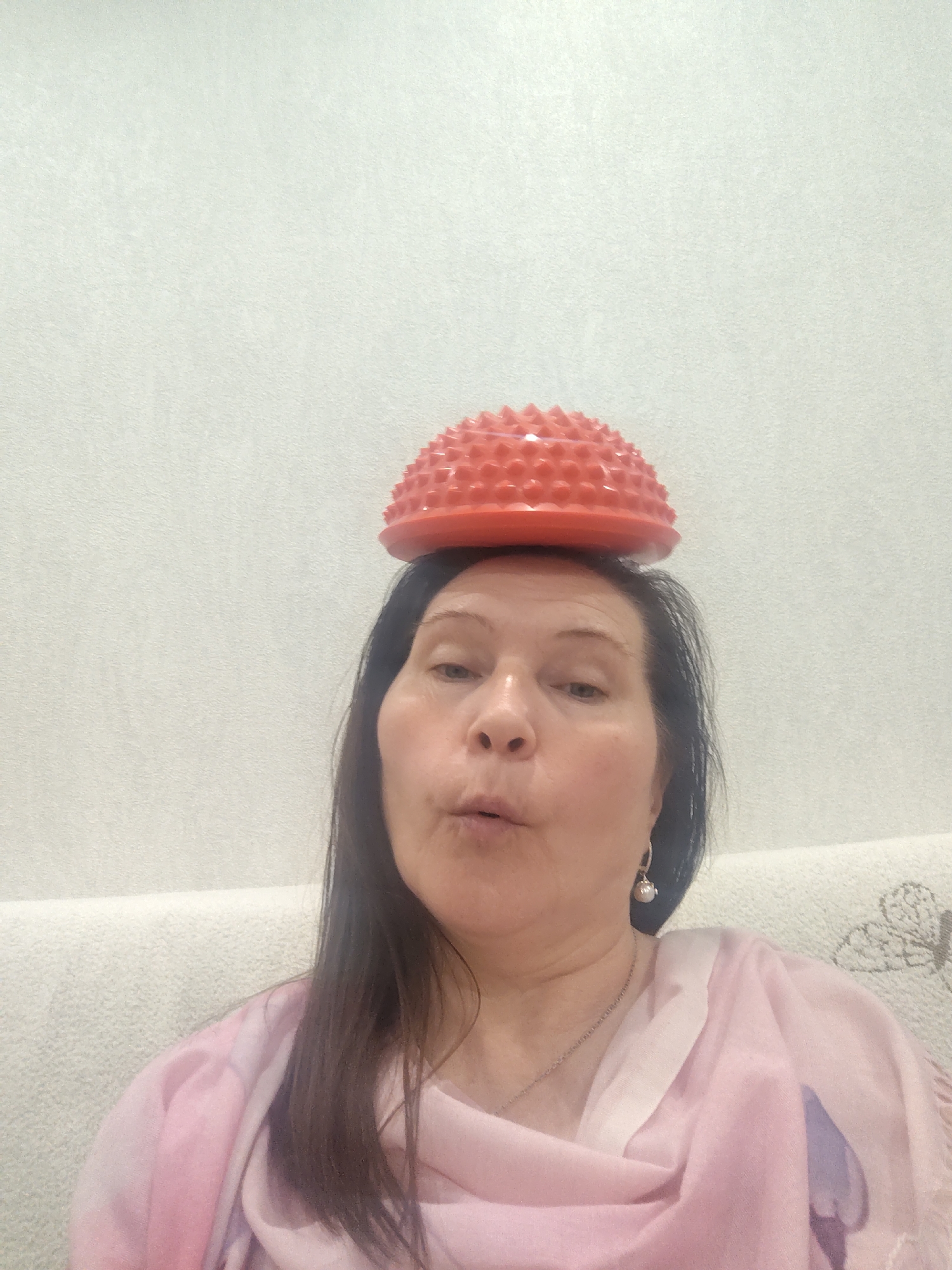A woman with dark hair poses indoors, holding a red, spiked dome-shaped object on her head. She has an expression of puckered lips. She is wearing a pink scarf, a silver necklace, and drop earrings. The background features a textured white wall and a light-colored sofa. The location is presumed to be Sarapul, Russia.