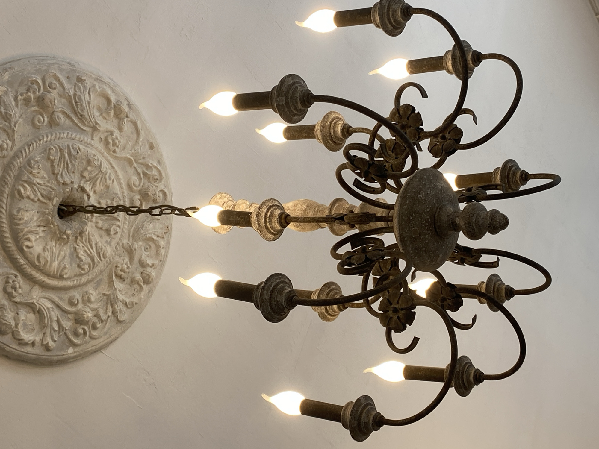 An ornate, rustic chandelier hangs from a white, textured ceiling. The chandelier has a central wooden element with carved details, from which metal arms extend, holding candle-shaped lights. The metalwork is dark and appears aged, with decorative floral elements. There are a total of eight visible lights, all illuminated and emitting a warm glow. Above the chandelier, a round, decorative ceiling medallion is visible, also with intricate relief patterns. The overall scene suggests an interior setting, possibly a dining room or entryway.
