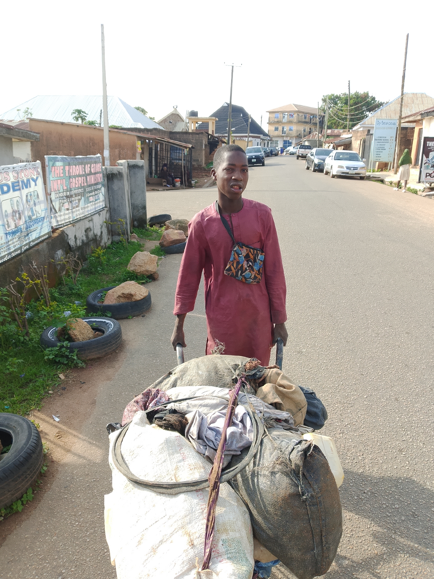 A young Black male, appearing to be in his early teens, stands on an asphalt road in what appears to be a Nigerian city. He is wearing a loose-fitting, dark red garment and has a colorful, small bag slung across his chest. He is pushing a large, overloaded handcart filled with various bundled items, including what appear to be textiles, sacks, and possibly plastic containers. The cart has two large wheels. The background shows a paved road with several parked cars and buildings lining the street. There are also signs posted on a wall to the left of the frame, one of which appears to be an advertisement for "THE THRONE OF GRACE INT'L GOSPEL MINISTRY." Several discarded tires filled with rocks are visible along the side of the road, suggesting this might be an area where such materials are collected or stored. The overall lighting suggests it is daytime, with clear skies.