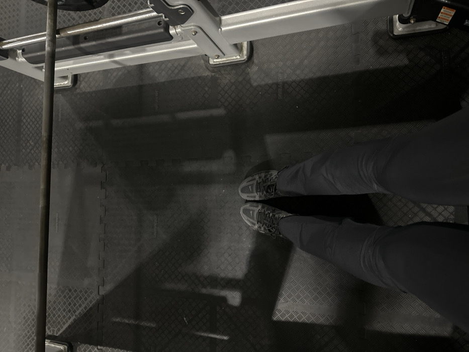 A top-down perspective captures a person standing on a black, diamond-patterned floor. The individual wears dark athletic shoes and dark pants. Above them, parts of a weightlifting rack are visible, including the metal barbell. Harsh lighting casts pronounced shadows across the floor, creating contrast and highlighting the texture of the surface. The overall tone is stark and functional, suggesting a gym setting.