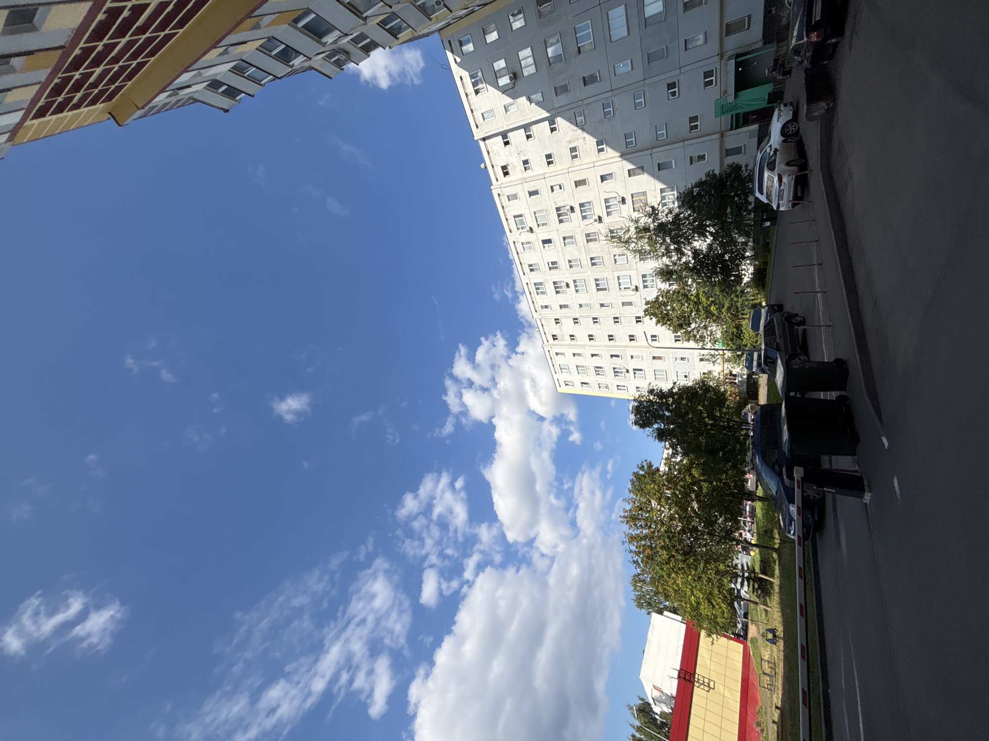 This is an outdoor, ground-level, wide-angle shot of a residential area in Naberezhnye Chelny, Russia, on a clear day with scattered white clouds. The scene is dominated by a large, multi-story apartment building with a gray facade and numerous windows. To the left of the apartment building, there are several deciduous trees with green and orange foliage, suggesting it is autumn. In the foreground, there is a paved road and sidewalk. Parked cars are visible on the road and in parking areas adjacent to the building and trees. A barrier gate is positioned across the road. Another building with a yellow and red facade is partially visible in the far left of the image. The perspective is slightly tilted, giving the impression of looking up towards the sky and the buildings.