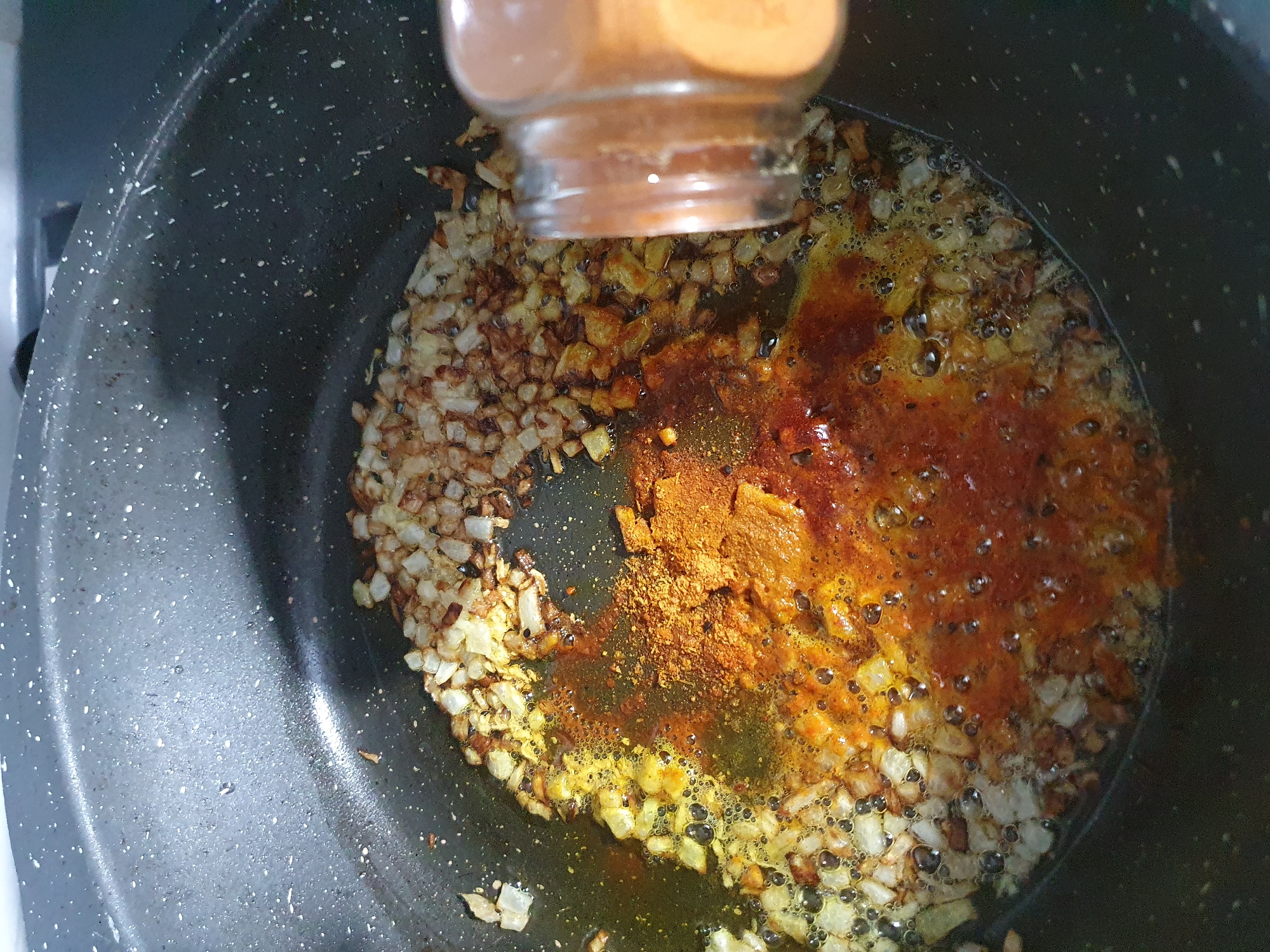 In a close-up shot of a dark, speckled pot, diced onions are being sautéed in oil. A spice blend, appearing to be a mix of red and yellow powders, is being added to the onions from a small glass jar held by an unseen hand. The spices are mixing with the oil and onions, creating a bubbling, vibrant mixture. The lighting is bright, highlighting the textures and colors of the food. The background is out of focus, showing the rim of the pot and a hint of a stovetop. The scene suggests a cooking process, likely the preparation of a flavorful dish.