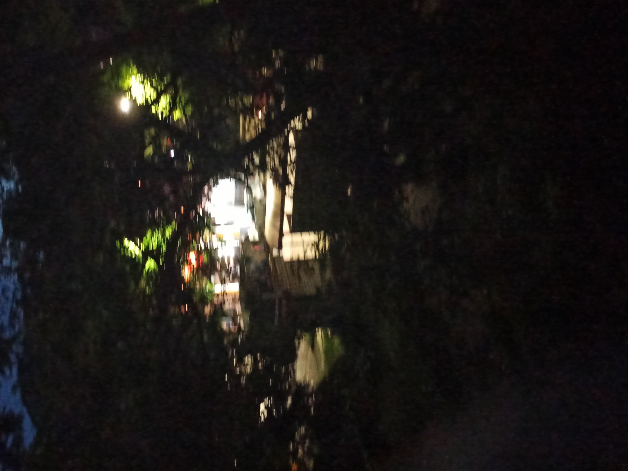 The image depicts a blurred, dark scene, potentially at night, in what appears to be an urban environment. On the left, there is a cluster of bright lights, possibly streetlights or lights from buildings, shining through trees. The building is reflecting on water, creating blurred light patterns. The right side of the image is obscured by darkness, likely due to shadows cast by trees or buildings. Overall, the photo captures a dimly lit urban setting with a focus on the contrast between light and shadow.