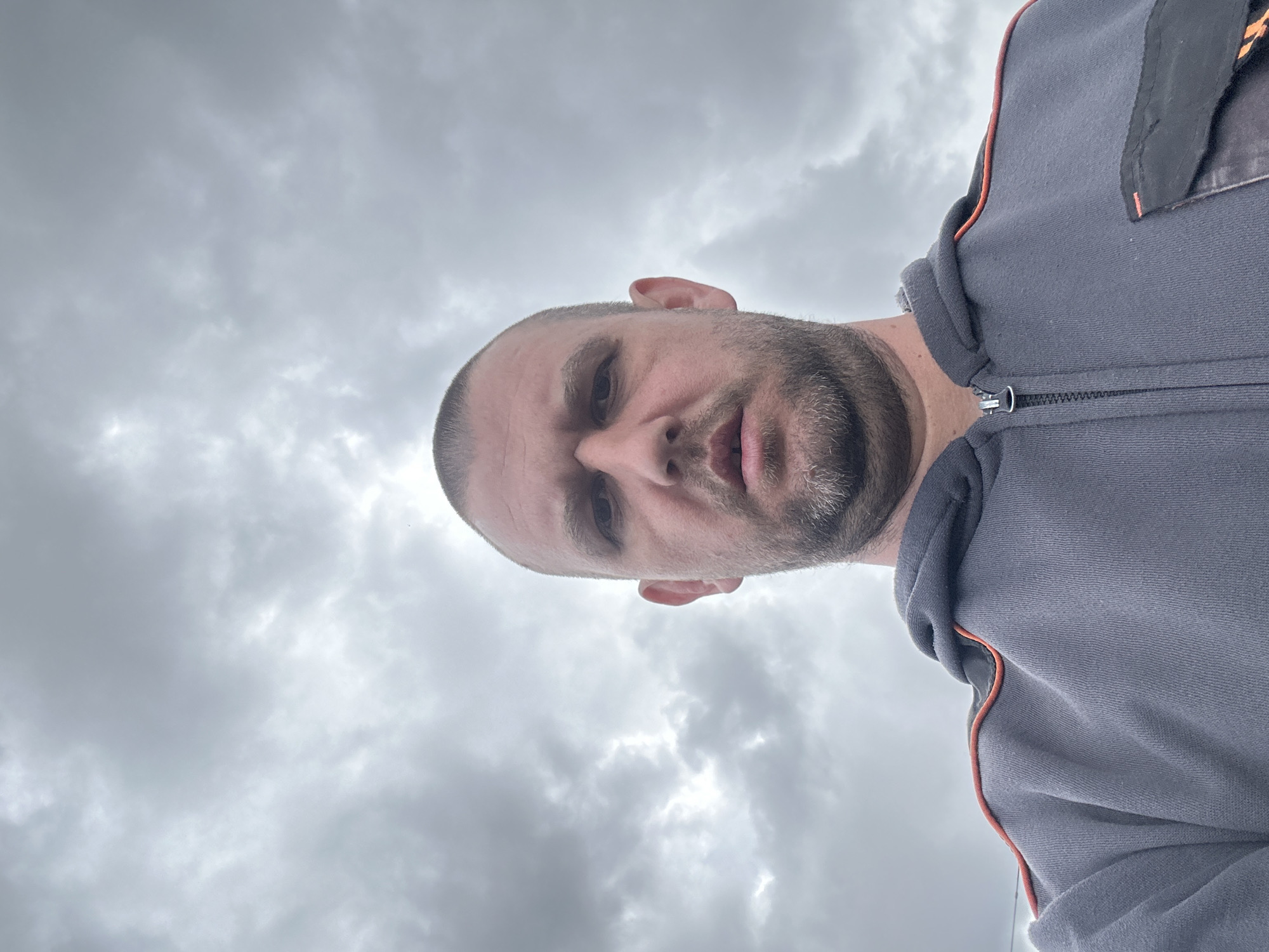 A man with a shaved head, light skin, and a short beard is looking down at the camera. He is wearing a grey zip-up hoodie with orange trim. The background is a cloudy, overcast sky. There is no information in the image to suggest the location is Sofia, Bulgaria.