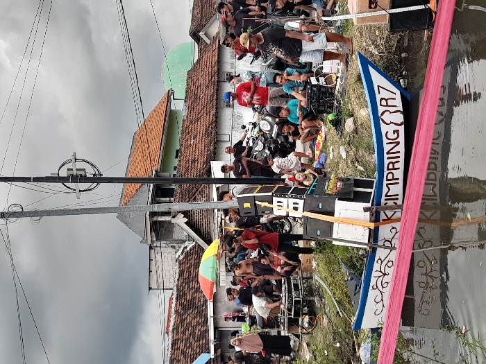 The scene depicts a group of people gathered near a canal in the Kecamatan Tanggulangin area of Indonesia. The canal is lined with green vegetation. A decorated boat with the words "CIMPRING R7" written on it, is partially submerged in the canal. The boat is laden with what appears to be speakers or musical equipment. The group of people consists of men, women, and children. Some are standing around, while others are seated. A woman in a hijab is standing near a blue motorbike. A man wearing a blue shirt is holding a microphone. There is a power pole in the center of the scene with power lines strung overhead. The sky is overcast with gray clouds. The scene is reminiscent of a community gathering.