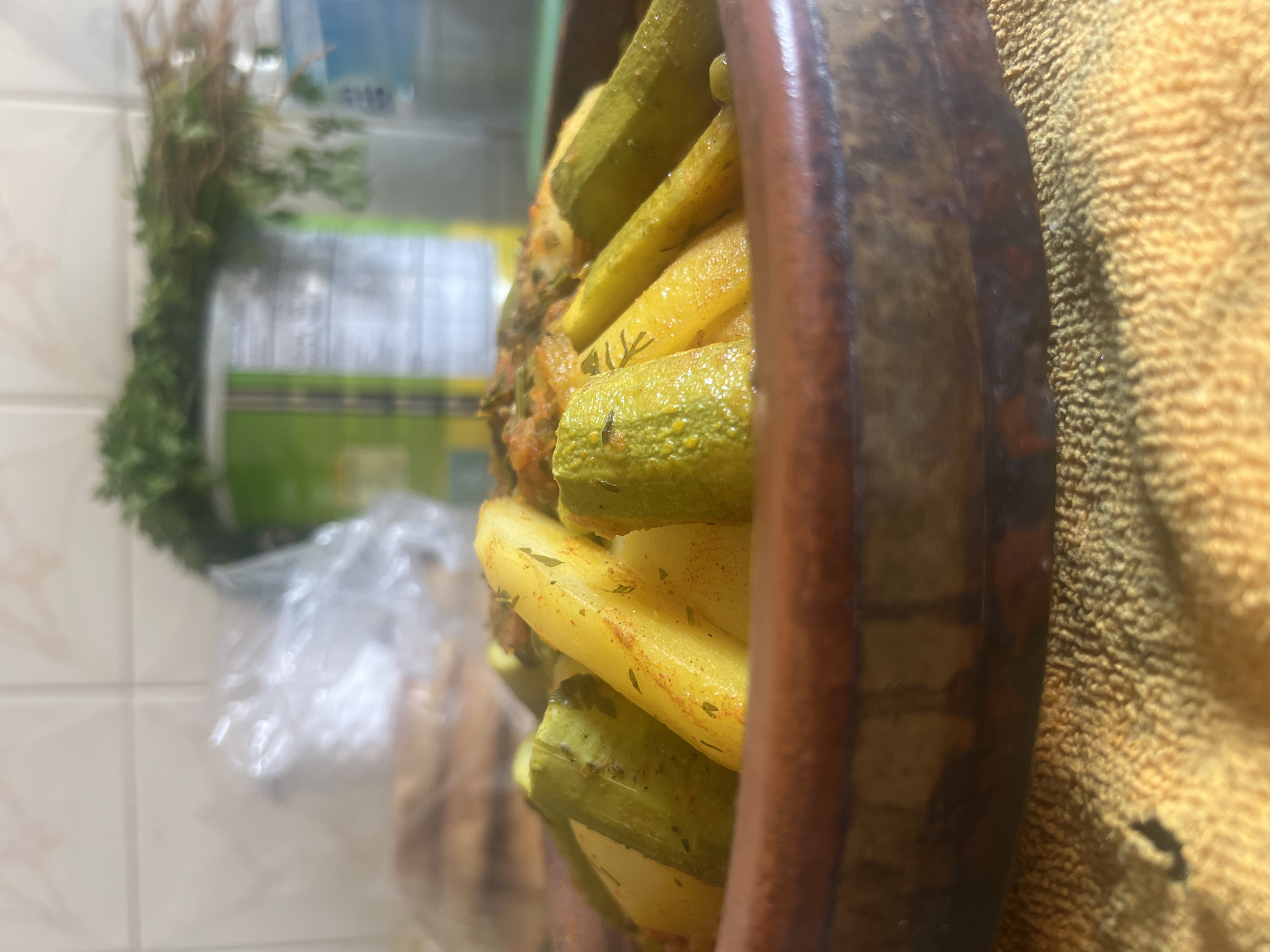 A close-up shot shows a tagine dish, containing cooked vegetables (potatoes and zucchini), beside a plant and a bag in what appears to be a domestic setting. The location, Ouaouizert, Morocco, is inferred from the prompt. The image offers no information about people, interactions, or specific actions.