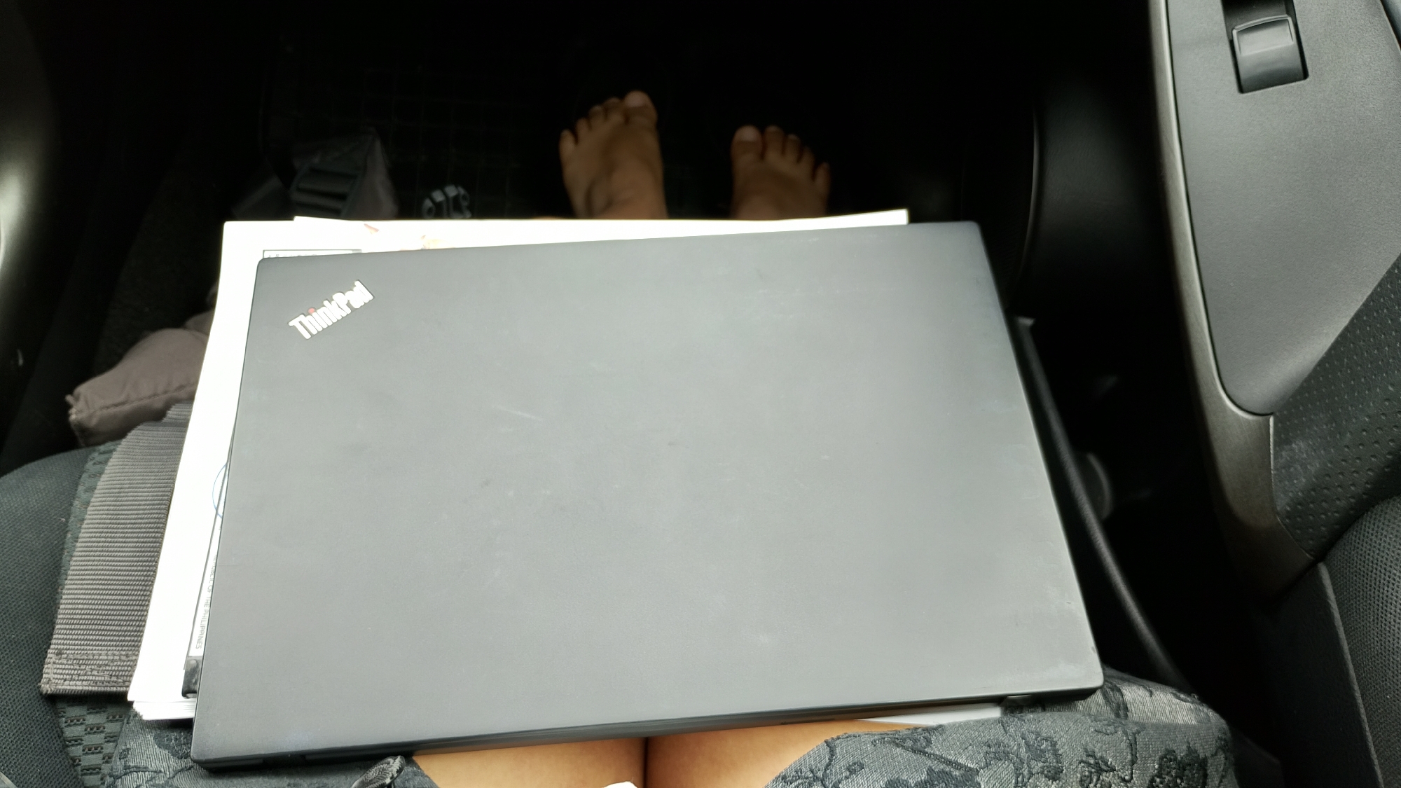An overhead shot captures a black Lenovo ThinkPad laptop placed on top of a stack of documents. The laptop is closed and positioned on a car seat. The driver's feet are visible behind the laptop, suggesting the car is parked. The interior of the car, including a portion of the dashboard, is visible in the background. The scene appears to be indoors, likely in a garage or parking lot in Sofia, Bulgaria, given the details in the image.