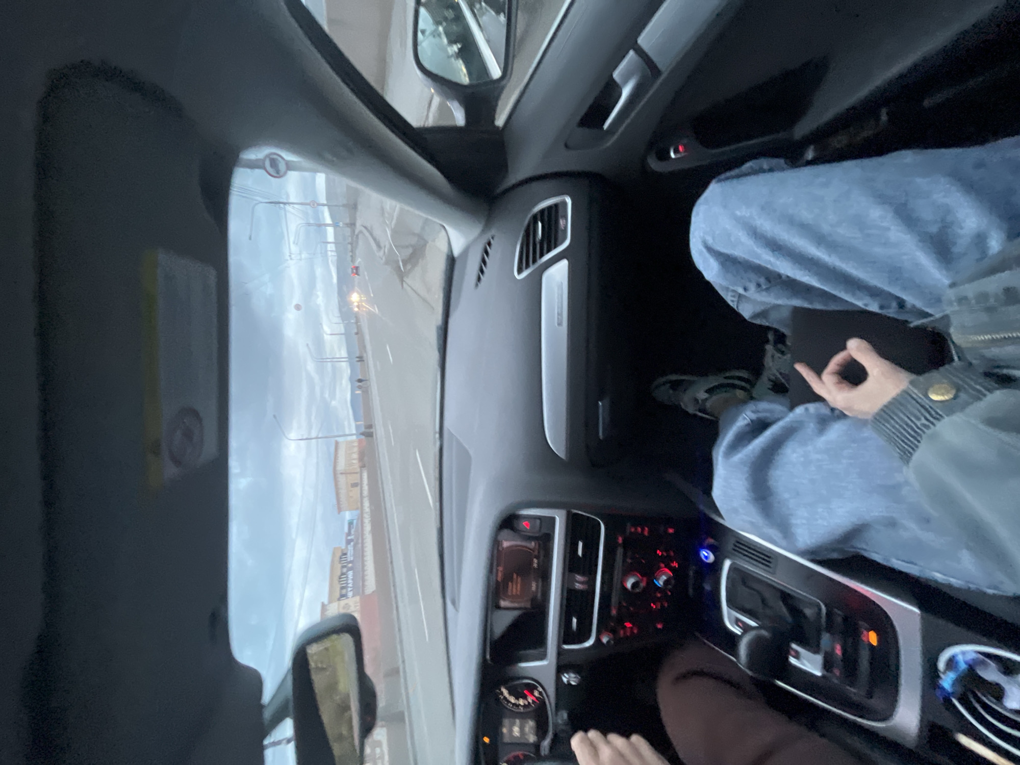 The image, taken from inside a car, shows a view through the windshield onto a road in Krasnoyarsk, Russia. The dashboard and part of the interior of the car are visible. A person wearing jeans and a jacket is in the passenger seat, making an "OK" hand gesture. The car's dashboard lights are illuminated, and the car's gear shift is visible. The environment outside shows a street with buildings, streetlights, and overcast sky.