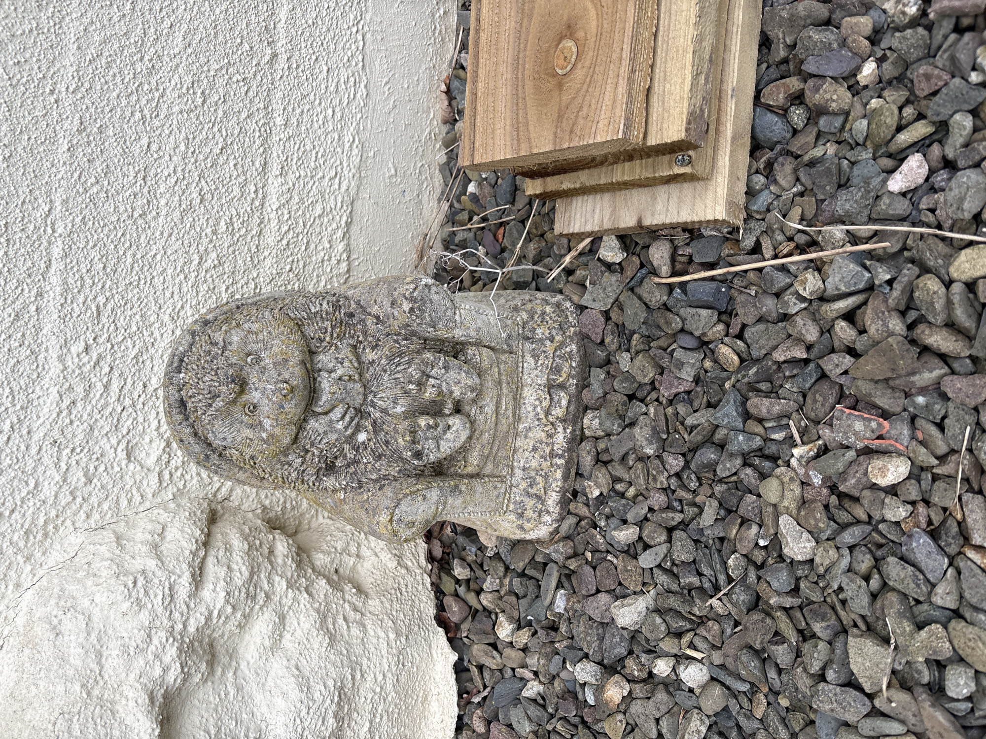 A stone Buddha statue sits adjacent to a white exterior wall and on a bed of gray gravel. Two pieces of light brown wood are placed near the gravel and the wall. The scene is outdoors, presumably in Longden, United Kingdom.