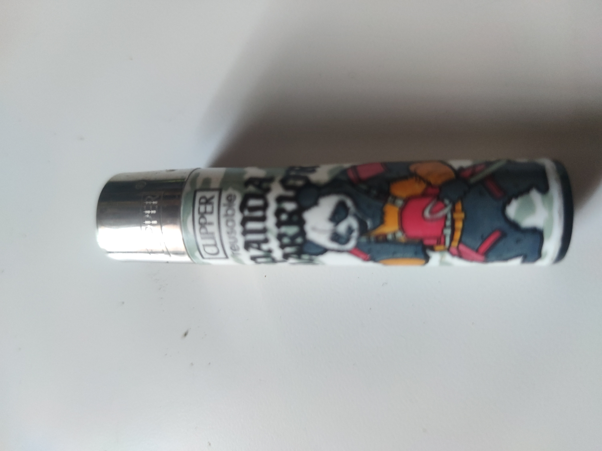 The image shows a Clipper brand lighter lying horizontally on a white surface. The lighter features a graphic of a panda dressed as a warrior. The brand name "CLIPPER" and the word "reusable" are visible on the lighter's body. The environment appears to be a plain, undecorated surface.