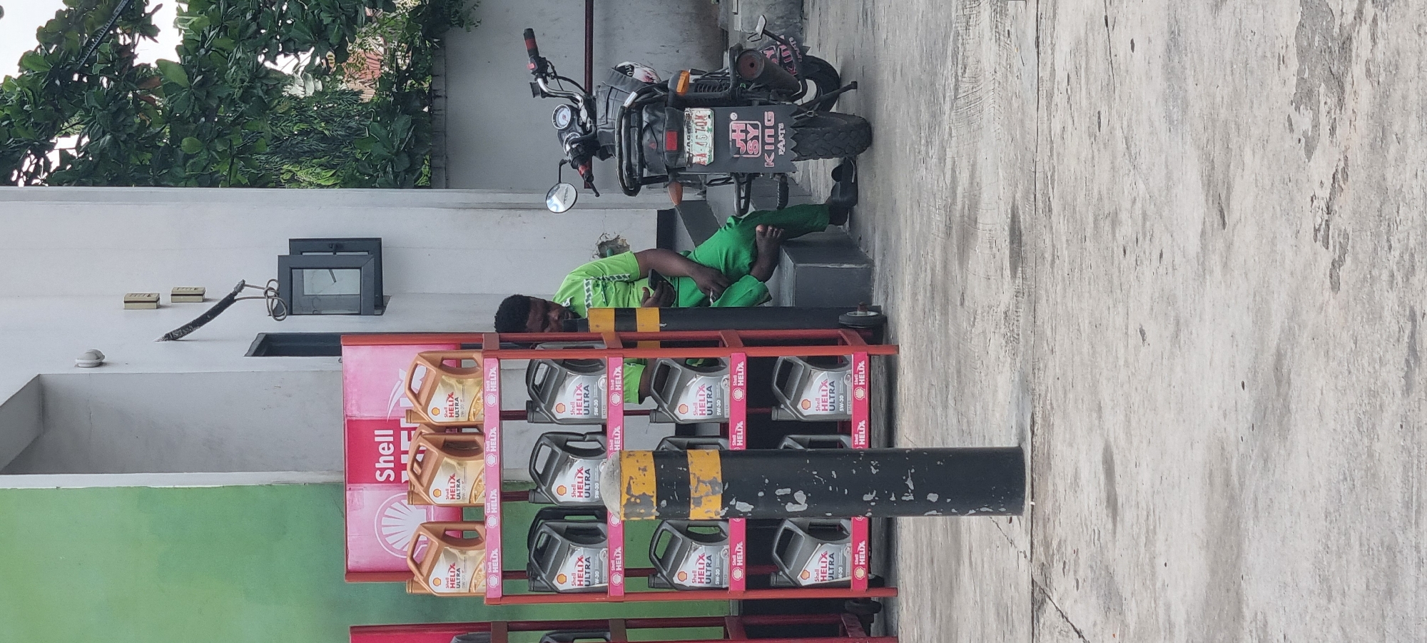 A man wearing a green shirt and green pants sits on a dark gray step to the right of a red shelf displaying Shell Helix Ultra motor oil bottles. He is looking at his phone, which he holds in his right hand. A motorcycle is parked to his right, with a license plate visible. The background includes a white building with a green wall to the left. The scene appears to be outdoors, possibly at a gas station or service center in Moba, Nigeria.