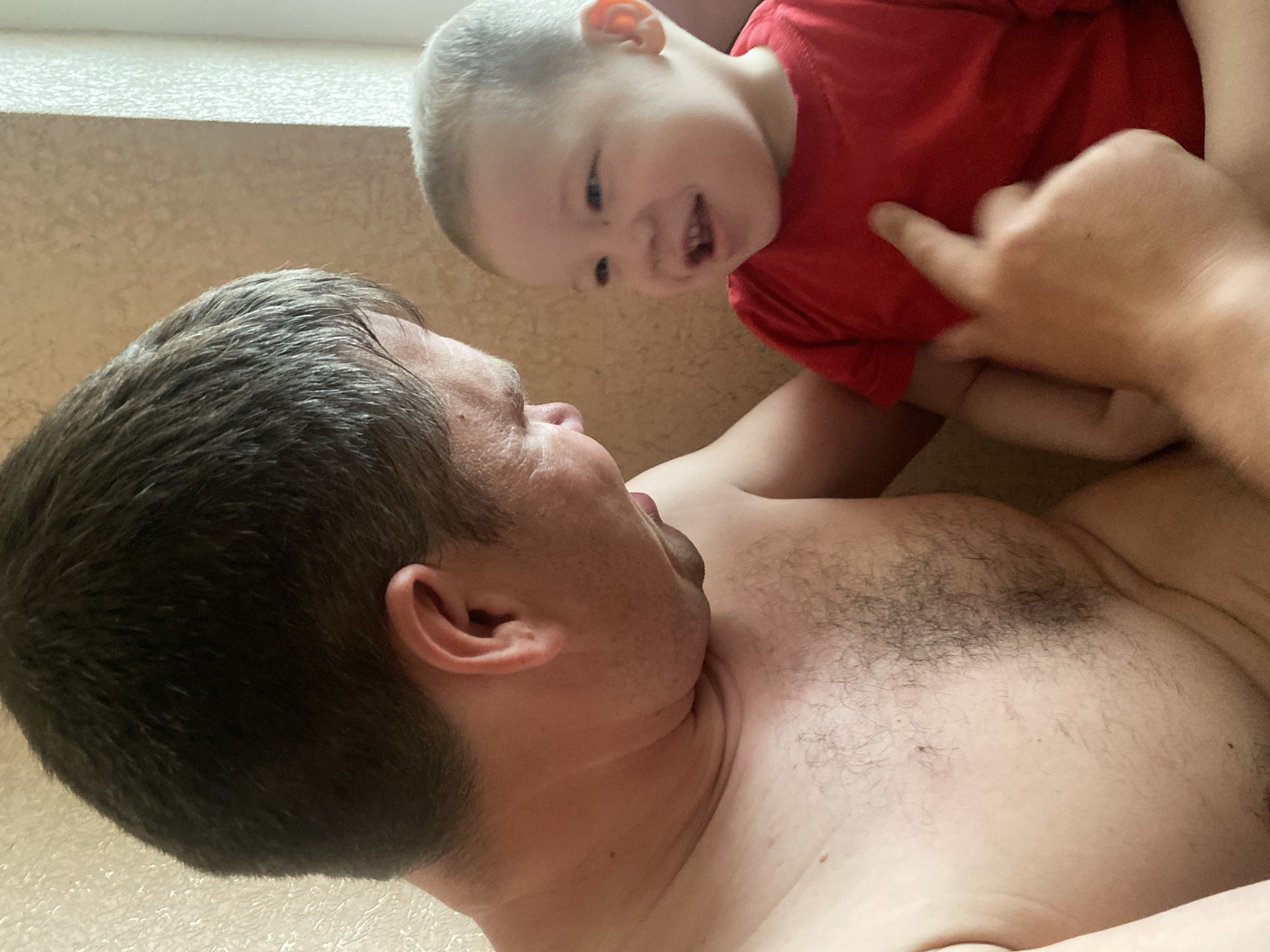 A man with dark hair is lying on his back. A young child in a red shirt is on top of him. The child is smiling and appears to be interacting with the man. The setting is indoors. The location is not evident from the image.