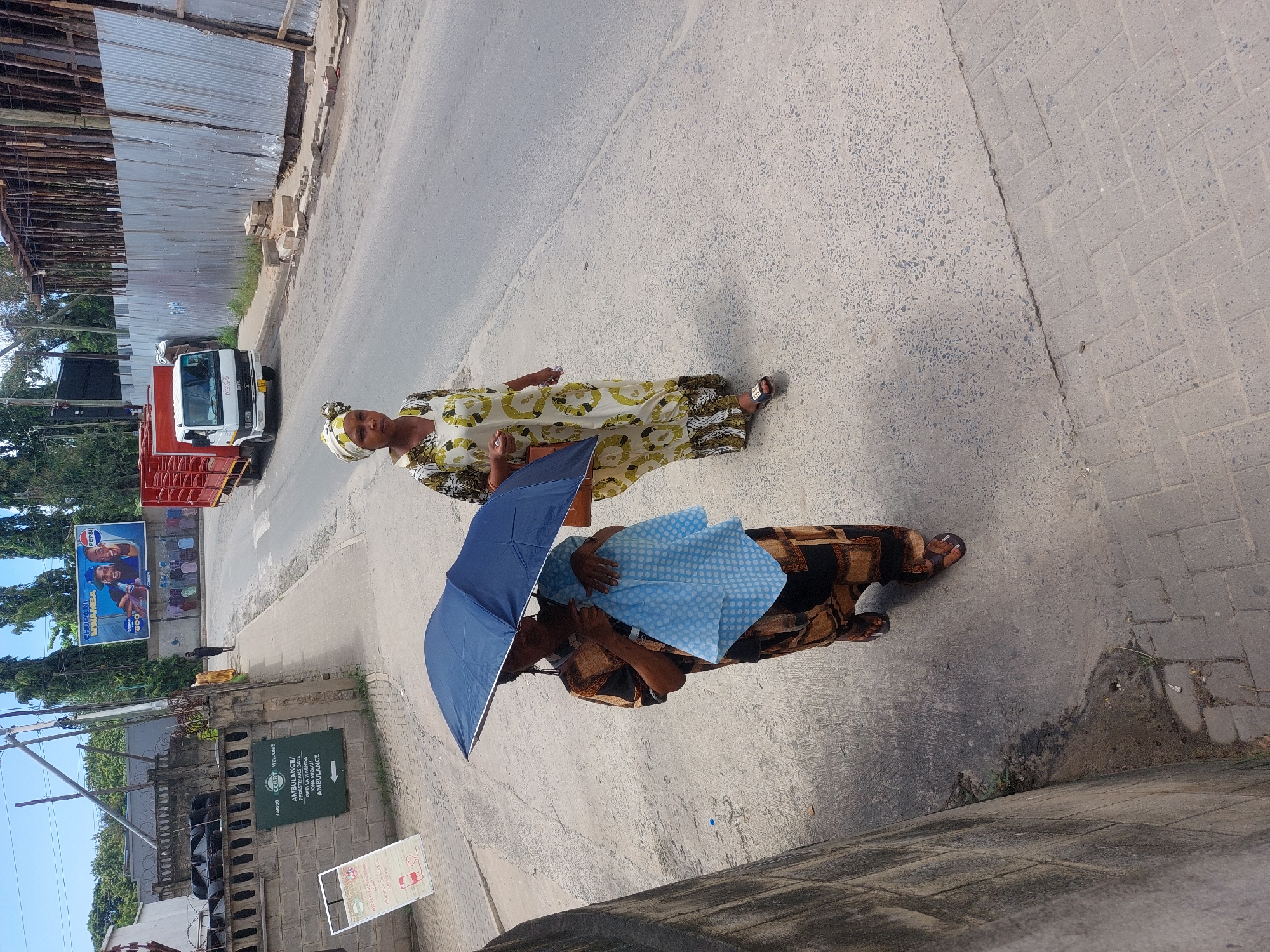 Two women are walking on a paved road. One is carrying a blue umbrella. The other wears a patterned yellow and white dress and a head wrap. A white truck with a red cargo bed is parked on the left side of the road. A building with a green sign that reads "AMBULANCE/PEDESTRIANS GATE GETI LA WAENDA KWA MIGUU/AMBULANCE" and the text "KARIBU" and "WELCOME" can be seen on the left. A billboard for Pepsi with the text "CHUPA NI MWAMBA" and "600" is also visible on the left. The scene appears to be in Dar es-Salaam, Tanzania, due to the Swahili text on the sign and billboard.