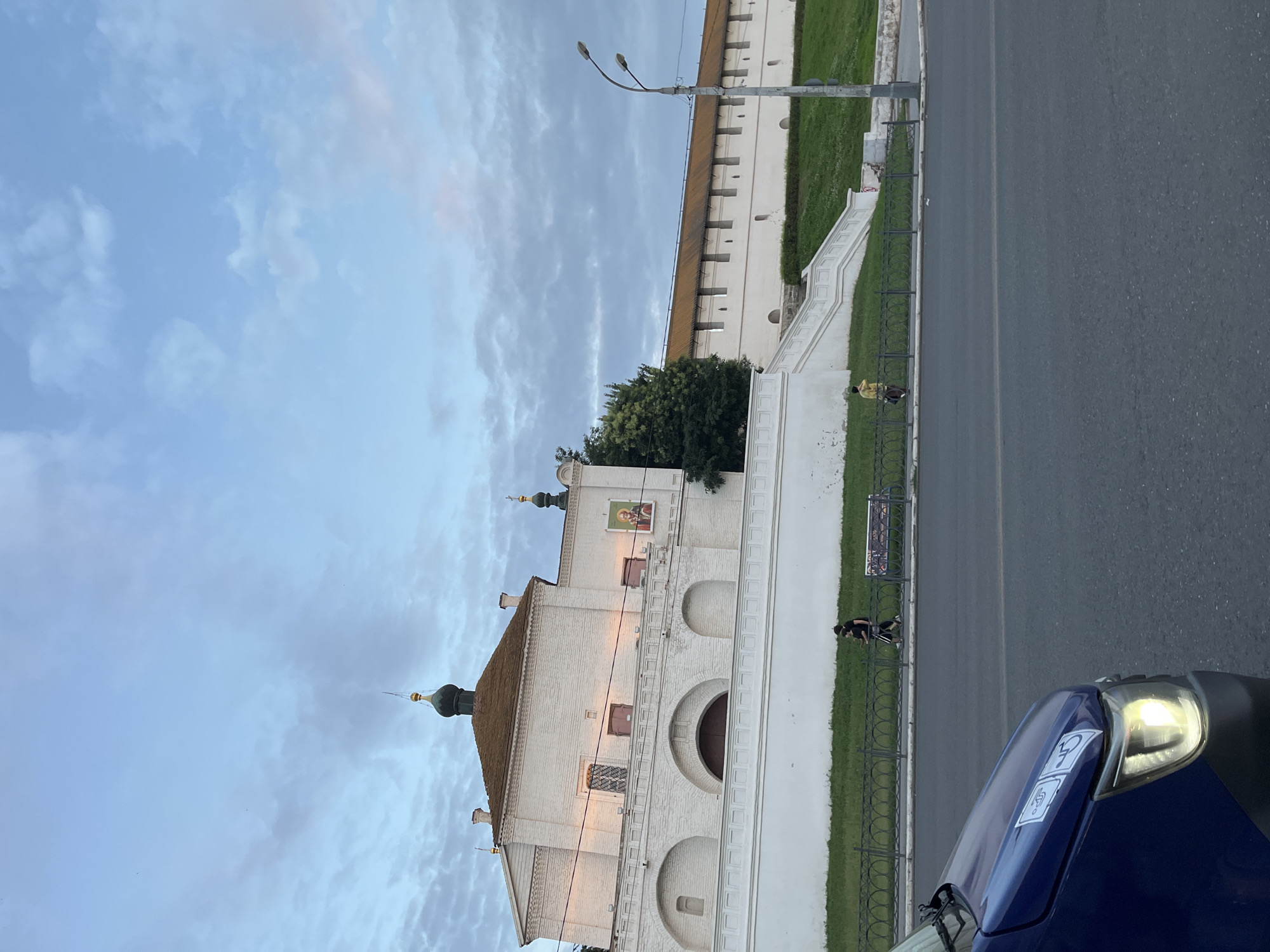 The image shows a wide view of a white brick fortress wall with a terracotta-colored roof in Astrakhan, Russia. The fortress has a prominent arched entrance and a decorative balcony. To the left of the fortress is a building with a green-domed cupola. In the foreground, a blue vehicle with its headlights on is parked on an asphalt road. A black metal fence separates the road from a grassy area, beyond which two individuals are walking. The sky is cloudy and appears to be either early morning or late evening.