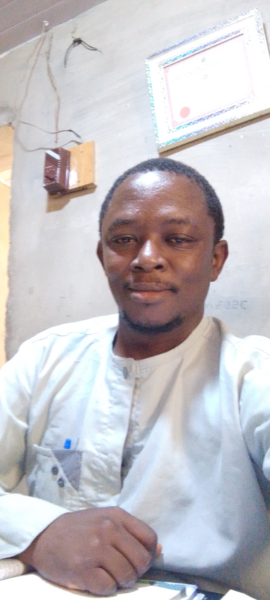 The image captures a man of dark complexion, likely in his 30s or 40s, looking directly at the camera with a calm and pleasant expression, featuring a slight smile. He has short, dark hair and a light beard. He is dressed in a light-colored, possibly white or off-white, traditional tunic-style shirt with a simple collar and buttons down the front. A blue pen is clipped into his left chest pocket. His right hand rests in the lower foreground, suggesting he is seated at a table or desk.
The background shows a plain, light-colored wall, typical of an indoor setting in Lokoja, Nigeria. On the upper left side, exposed dark electrical wires run down the wall, leading to a small, dark brown electrical box mounted on a wooden base. In the upper right, a framed document, likely a certificate, hangs on the wall. The details within the frame are indistinct due to blur and brightness, though a red circular seal is faintly visible. Some faint dark writing, resembling "FO226," is also visible on the wall to the right of the man's head. The lighting is consistent with an indoor environment.