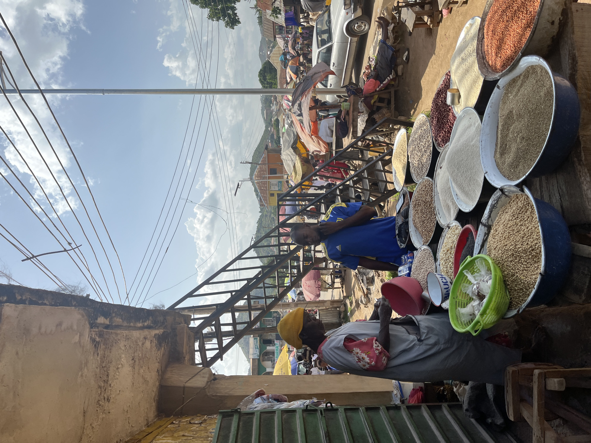 This image depicts a vibrant outdoor market scene in Gwong, Nigeria. The primary subjects are people and various food items displayed in large bowls and baskets. A person wearing a yellow head covering and a grey shirt with a floral pattern is seated at a stall, interacting with their wares. Another person in a blue and yellow t-shirt stands nearby, possibly a vendor or shopper. The market is set up along a dusty road with a backdrop of buildings and a gently sloping landscape, suggesting a hilly terrain.
Numerous large bowls are filled with different types of grains, legumes, and seeds, including what appears to be rice, beans, and peanuts. A bright green basket holds smaller plastic bags of goods. A red bucket is also visible. In the background, a silver car is parked, and several market stalls with colorful awnings and umbrellas are set up. A metal staircase leads up to an upper level of the market or an adjacent building.
The time of day appears to be daytime, with bright sunlight casting shadows and illuminating the scene. The sky is blue with scattered white clouds. The atmosphere seems busy and bustling, typical of a local market. Visible text includes "PHARMACY" on a sign in the background. The overall impression is one of lively commerce and community activity.