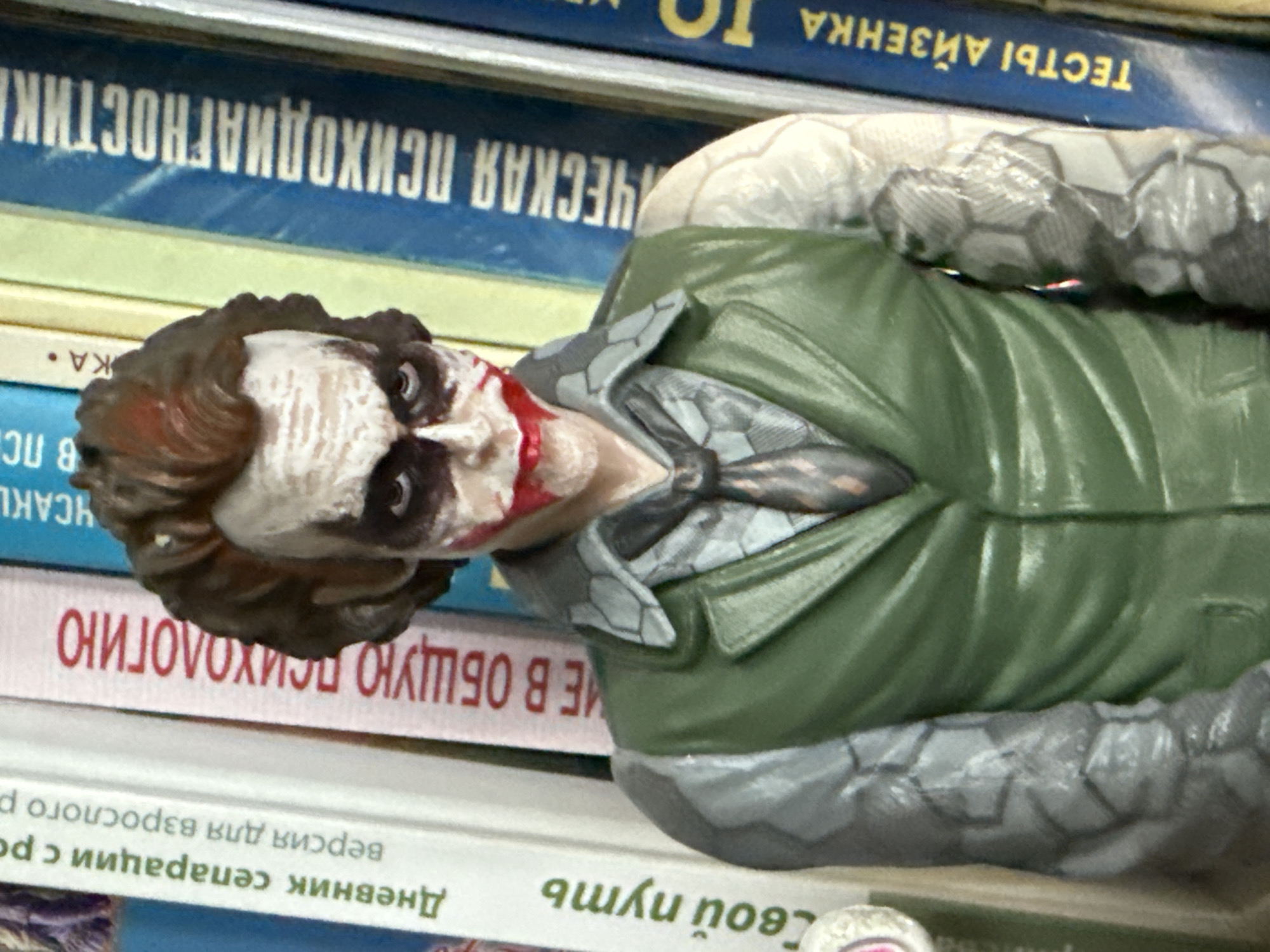 The image is a close-up, eye-level, medium shot of a Joker figurine positioned in front of several books. The figurine, which is the primary subject, is detailed and depicts the character with white face paint, dark eye makeup, and red lipstick, a common portrayal of the Joker. It is wearing a green vest over a grey patterned shirt and tie. The figurine's arms are visible and appear to have a textured, almost reptilian pattern.
The background consists of a stack of books, vertically aligned. The spines of these books display text in Russian. Some visible text includes "Дневник сепарации с р", "версия для взрослого р", "свой путь", "ПСИХОЛОГИЮ", "ЧЕСКАЯ ПСИХОДИАГНОСТИКА", and "ТЕСТЫ АЙЗЕНКА". The presence of these specific book titles and the overall context suggest this scene might be in a personal collection or a library setting.
There is no indication of the city of Simferopol, Ukraine, or any specific location within it. The image captures a still, posed subject without any dynamic action or interaction. The lighting appears to be natural or from an overhead source, illuminating the figurine and the books evenly.