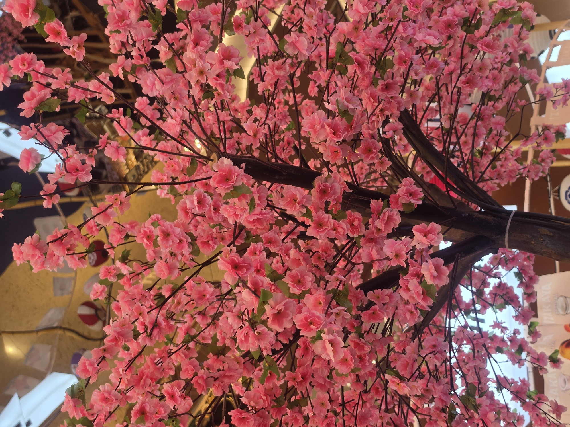 The image shows a close-up view of a flowering tree with numerous pink blossoms. The tree's branches are dark brown and visible amidst the dense flowers. The background is a mix of light and shadow, with hints of other decorative elements. The flowers dominate the composition, creating a visually lush scene. Based on the decorative elements, it can be speculated that the location is the city of Bangkok, Thailand, and potentially an indoor setting with artistic lighting.
