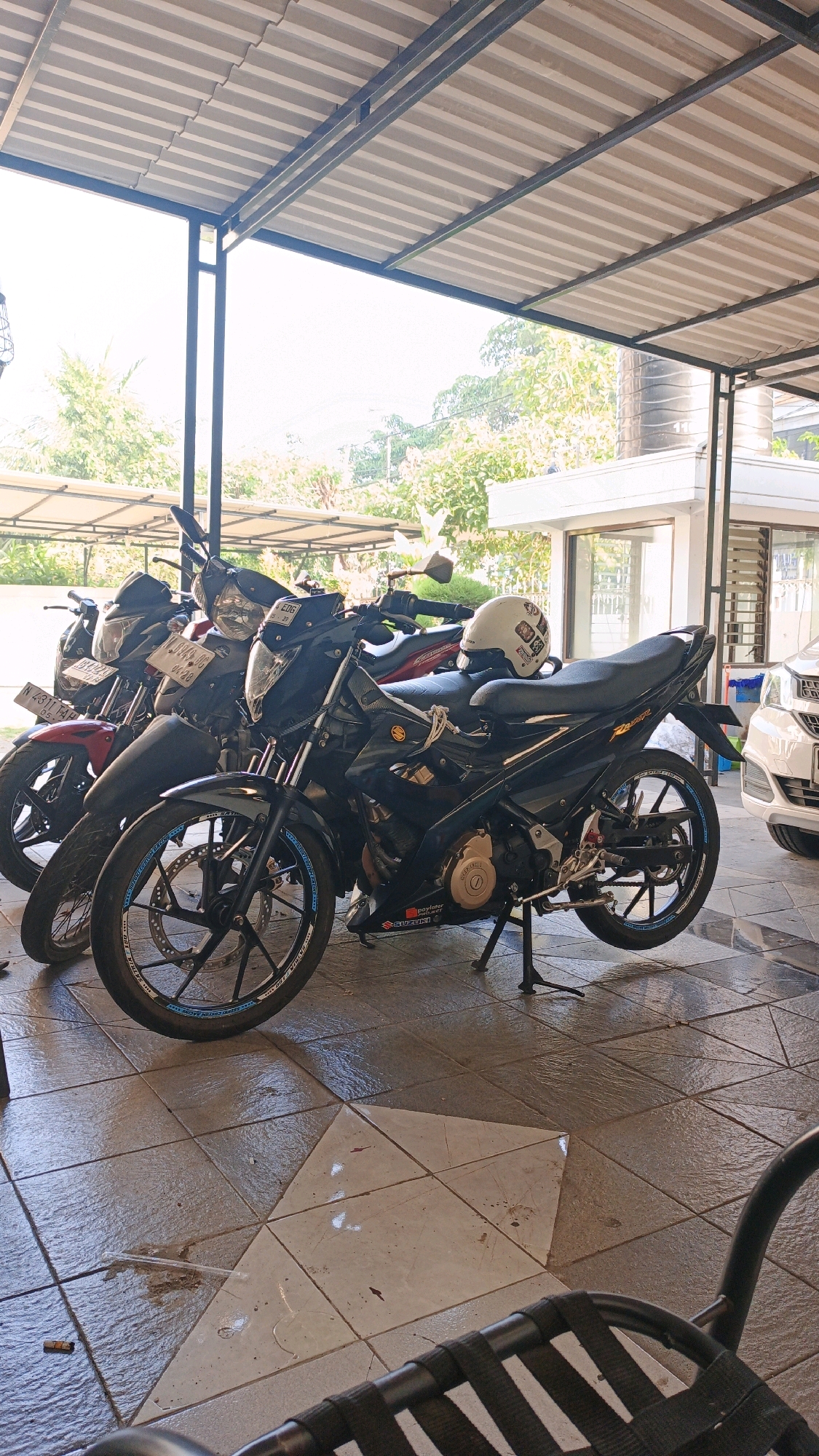 The image shows a row of parked motorcycles under a covered patio area with a corrugated metal roof. The motorcycles are parked on a tiled floor, some of which appears to be wet, with a small puddle and some liquid trails visible. In the foreground, a portion of a black metal chair is visible. The motorcycles are of varying colors, including black, red, and one with a white helmet on its seat. The background reveals lush green trees and foliage, suggesting an outdoor environment, possibly a residential or commercial property in a tropical or subtropical climate. A white car is partially visible on the right side of the image. The overall scene depicts a typical urban setting in Surabaya, Indonesia, likely a parking area or a residential driveway. There are no people or animals visible in the image.