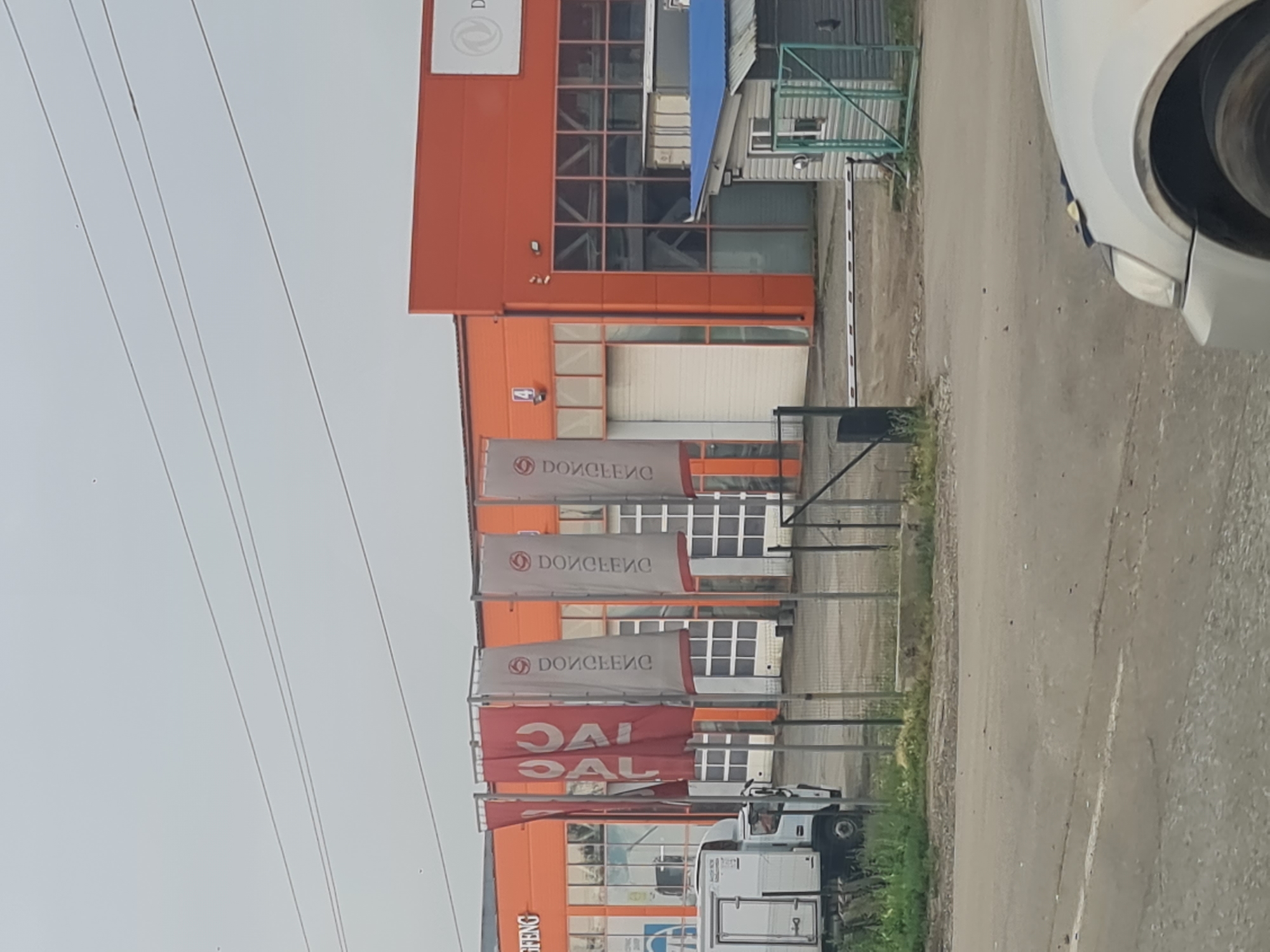 The image shows an orange building with a lot of windows and some flags in front of it. There is also a white truck and a silver car. The building appears to be a business or commercial property. It is difficult to ascertain the exact location from the image alone. However, based on the flags which may represent brands and the overall architecture, it is likely an industrial or commercial area. There are no people visible in the image.