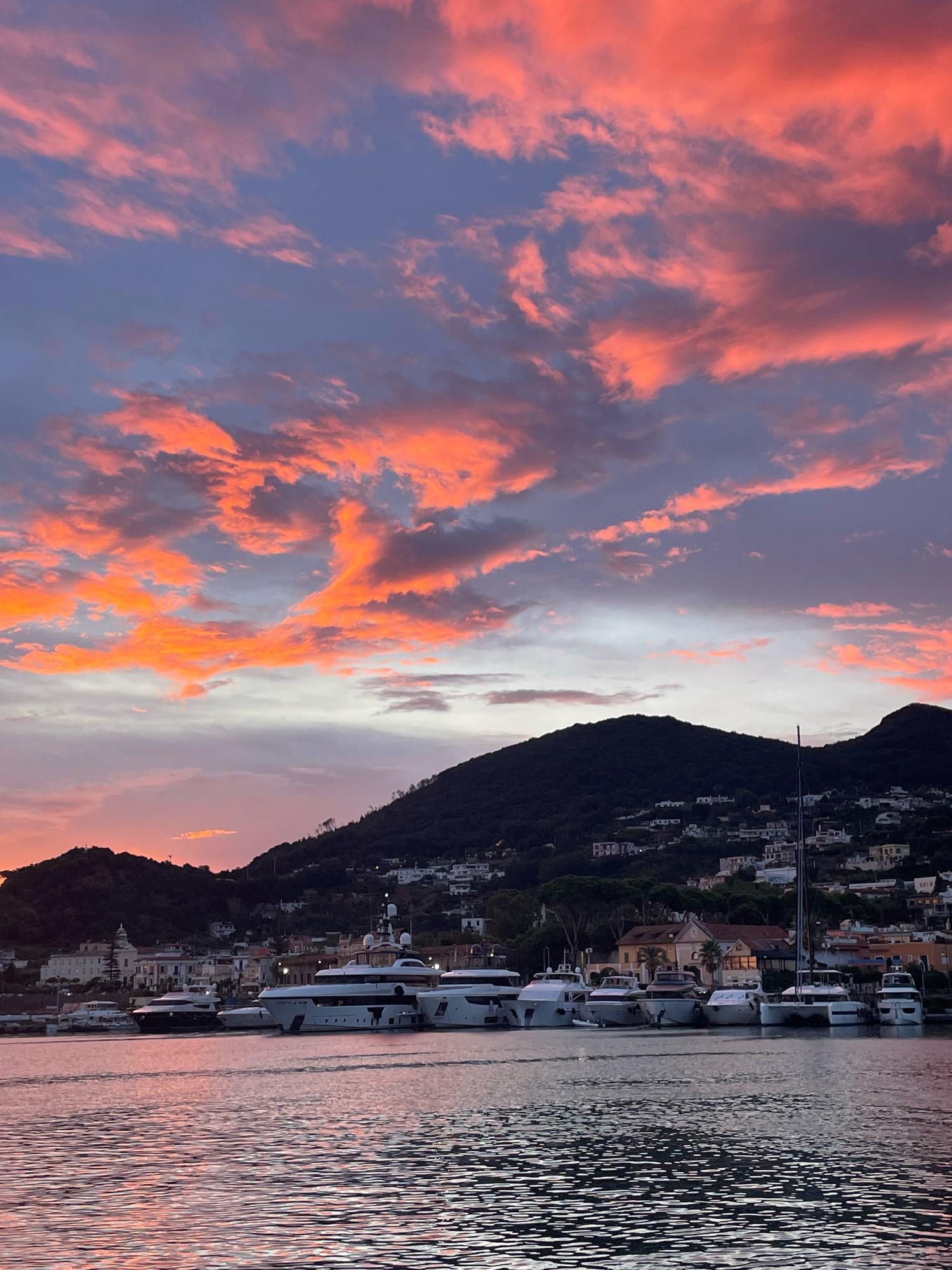 This is a vertical, eye-level, full shot of a harbor at sunset in Bagni, Italy. The sky is filled with dramatic, vibrant clouds in shades of orange, pink, and purple, streaked against a dusky blue. The water in the foreground reflects the colors of the sky. Lined up along the harbor are several large white yachts and a few smaller boats. In the background, a lush green hillside rises, dotted with white buildings and trees, suggesting a Mediterranean coastal town. The scene is peaceful and picturesque.