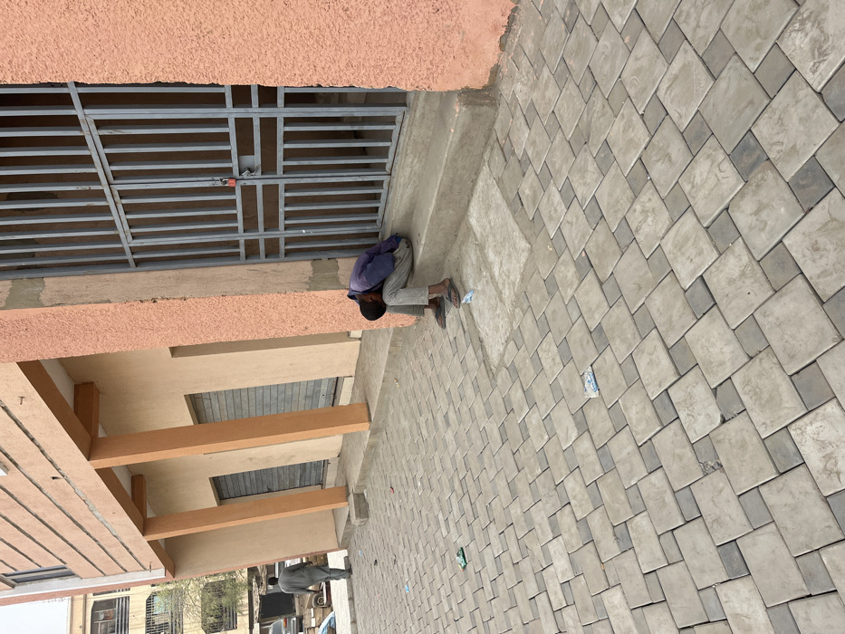This image depicts an outdoor urban environment in Sabon Gari East, Nigeria. In the foreground-right, a male individual is seated on a concrete step against a building featuring a peach-colored stucco facade. He is dressed in a dark blue long-sleeved shirt, light grey trousers, and flip-flops, with his head bowed towards his knees. Adjacent to him is a grey metal security gate secured with a red padlock. Two light-colored wrappers are present on the step near his feet. The ground is covered with interlocking grey concrete pavers, upon which several pieces of litter, including green and white plastic wrappers, are scattered. To the mid-left, a second male individual, wearing a light grey long garment, walks away from the viewer. Behind this individual, a beige building with three prominent light orange columns and three closed storefronts with metal roll-down shutters is visible. In the background, further urban structures and partially visible vehicles indicate a street setting. The scene is under bright daylight.