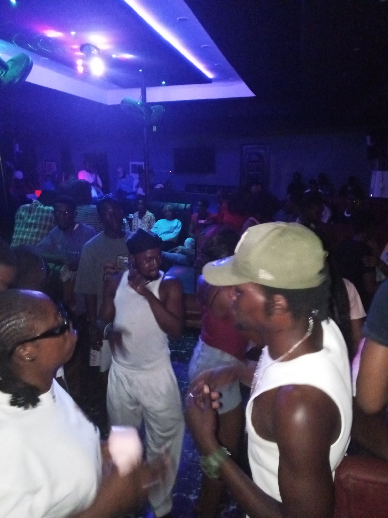 A crowded nightclub interior in Lokoja, Nigeria. Dimly lit with blue lighting and additional disco lighting on the ceiling. Various people are present, with some seated in the background and others standing and interacting in the foreground. In the foreground, there are several individuals wearing casual clothing, including a man in a white tank top and gray sweatpants, a woman in a white shirt and braids wearing glasses, and a man in a white tank top and a green baseball cap. The crowd appears to be engaged in socializing and dancing. There is a pole covered in faux foliage in the background.
