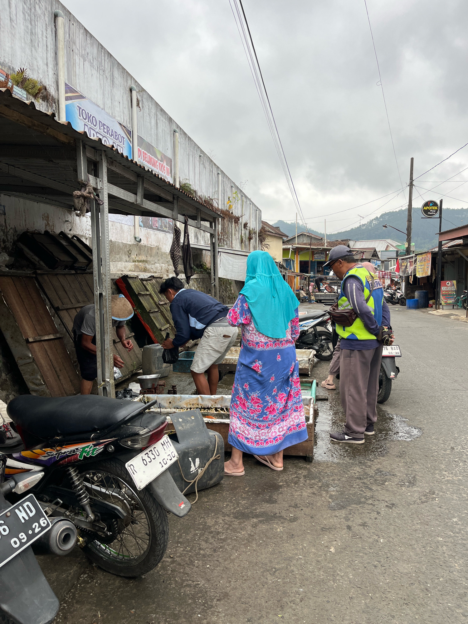 In Karangkobar, Indonesia, on a cloudy day, a street scene unfolds with several individuals and parked motorcycles. In the foreground, a black motorcycle is parked on the wet asphalt. To its right, two men are actively engaged in an activity beneath a weathered awning of a building that features a sign in Indonesian reading "TOKO PERABOT". One man, wearing a blue long-sleeved shirt and light-colored shorts, appears to be handling a plastic bag. The other man, wearing a grey t-shirt and a hat, is partially obscured, leaning into a wooden structure.
Further down the street, a woman wearing a bright blue hijab and a colorful floral dress is standing. Beside her, a man in a high-visibility yellow vest over a blue shirt and grey pants is facing away from the camera, seemingly interacting with something near a parked motorcycle. Another person, possibly a woman, is partially visible behind him. The street is lined with buildings and appears to be a commercial area. A sign for "APOTEK" (pharmacy) is visible in the distance. The overall impression is of a bustling local market or commercial street.