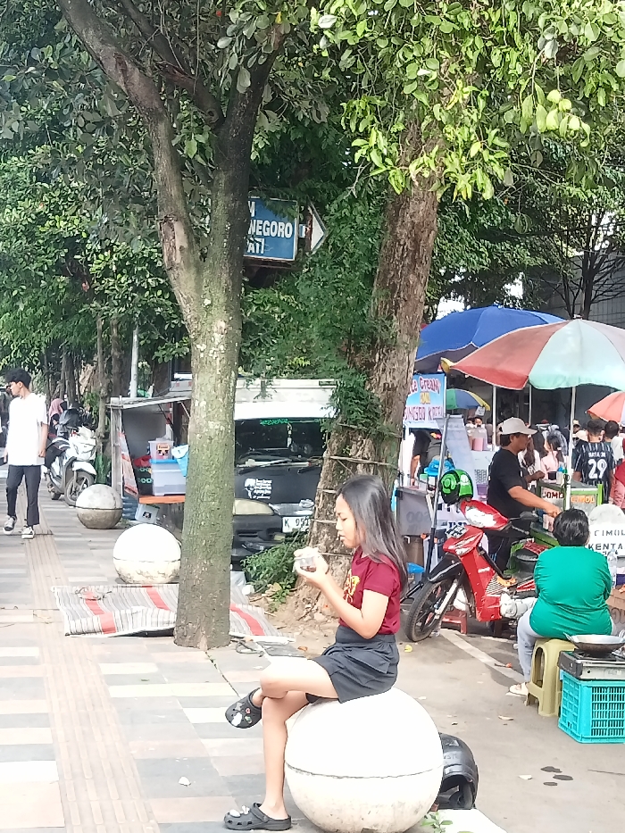 A young girl in a maroon shirt and black shorts sits on a large, white spherical bollard on a tiled sidewalk. She's focused on something in her hands. Behind her, street vendors and motorcycles are visible. A blue sign with white writing points to "NEGORO". Trees line the street, providing partial shade. A man in a white shirt and black pants walks on the left sidewalk. Other people are visible in the background near the vendor stalls.