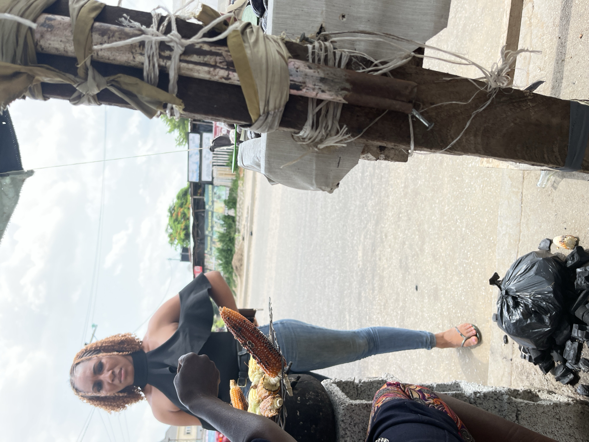 A scene in Abuja, Nigeria depicts a woman standing by a roadside corn vendor. The woman is wearing a black top and blue jeans, with silver flip-flops. The vendor has a charcoal grill with corn on it, and there is a black trash bag with charcoal next to the grill. The street and buildings are visible in the background.