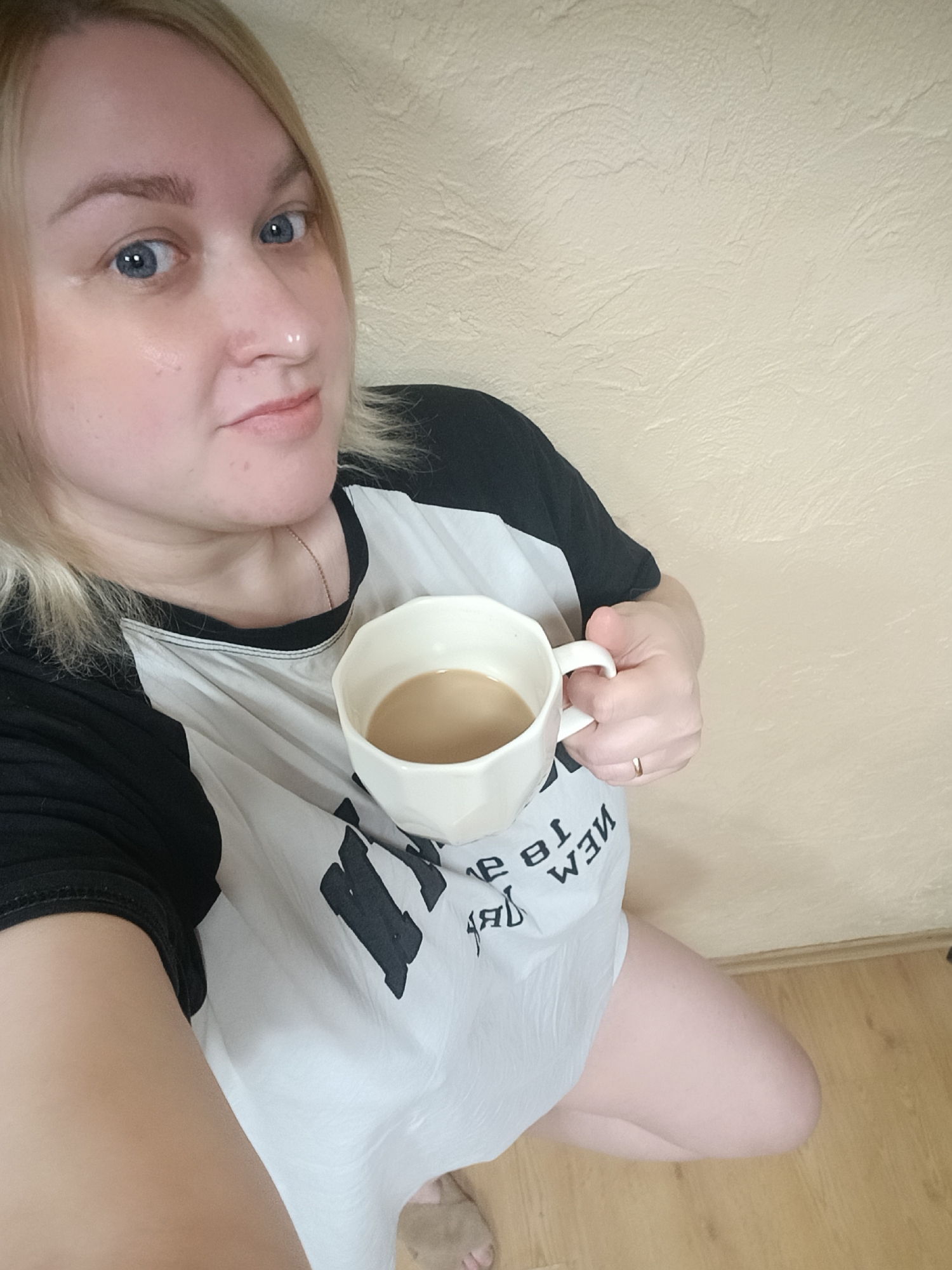 An indoor selfie features a woman, presumably in Chaikovsky, Russia, looking directly at the camera with a calm, subtle smile. She has blonde hair and striking blue eyes. Her fair skin shows some minor imperfections. She is wearing a white raglan-style t-shirt with black sleeves. The front of the shirt displays mirrored black text and design, with parts like "NEW 18" and "ЯЗ МЭМ" (likely "MEM" or "MOM" reversed) visible.
In her right hand, she holds a white, faceted ceramic mug containing a light brown beverage, likely coffee or tea with milk. A thin gold ring is visible on her right ring finger. She appears to be in a kneeling or squatting position, with her bare right leg and knee visible. A glimpse of her left foot, possibly in a light-colored slipper, can be seen at the bottom left.
The background consists of a textured, light beige or off-white wall. The lower portion of the image reveals a light-colored wooden or laminate floor. The lighting suggests it is daytime.