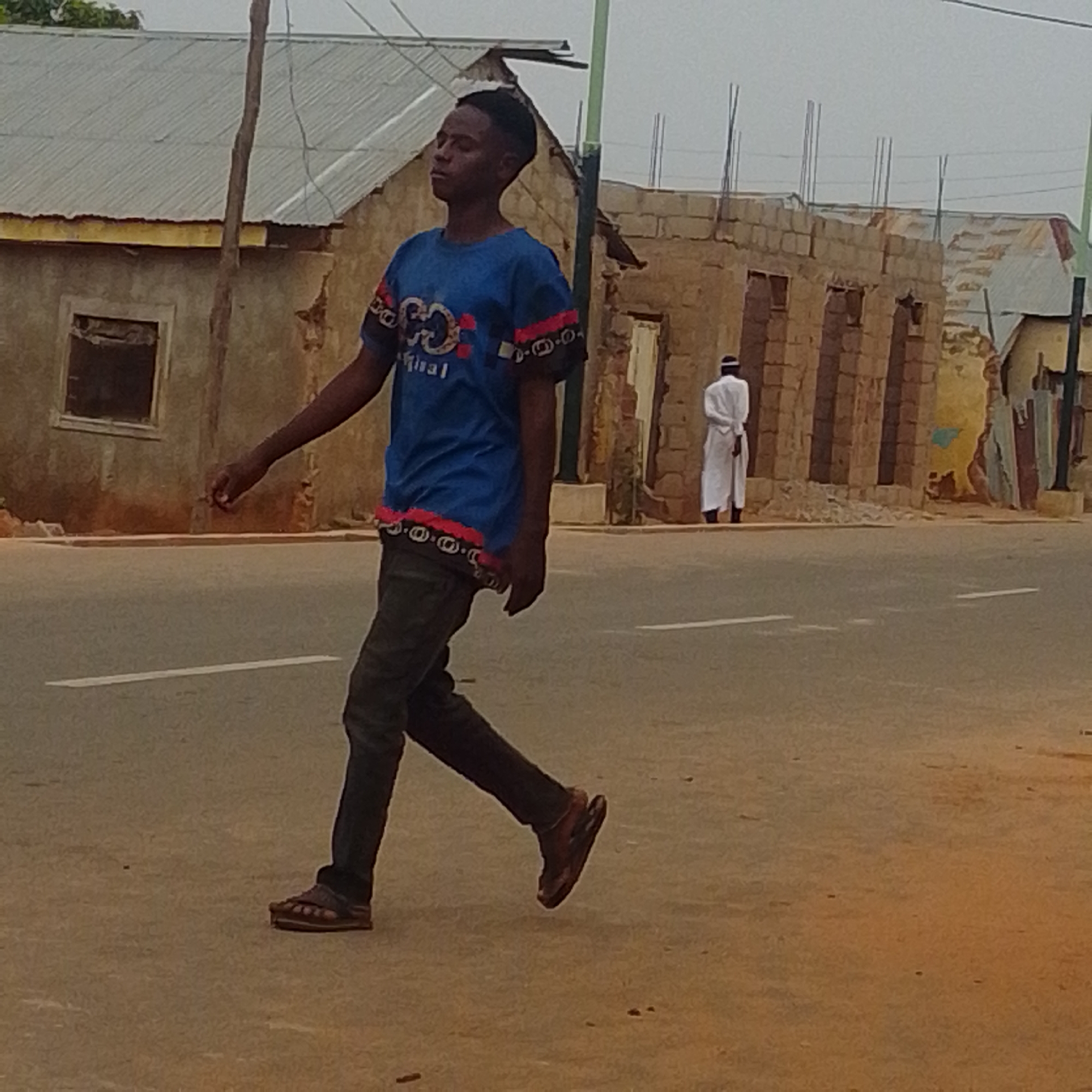 A young male individual wearing a blue t-shirt with a red and black patterned trim and dark pants is walking across a paved road in Tempure, Nigeria. He is wearing dark sandals. In the background, an older male in a white robe and cap stands near unfinished brick buildings, suggesting construction or disrepair in the area. The environment appears to be an urban setting with a cloudy sky overhead.