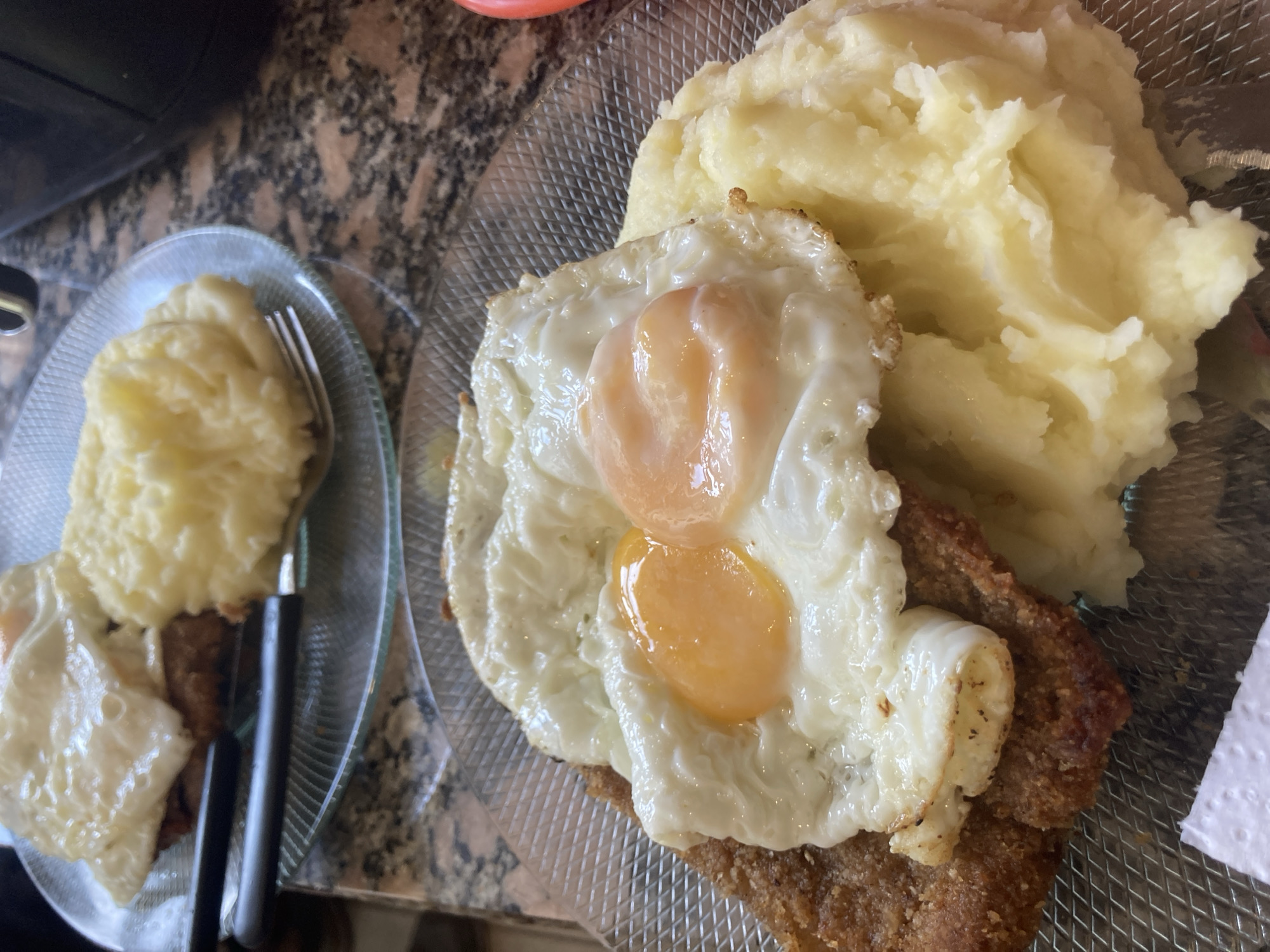 The image shows two plates of food. Each plate contains a portion of mashed potatoes, a breaded cutlet, and a fried egg with two yolks. The plates are on a surface that appears to be a kitchen countertop. There is a fork on the left plate. The location is not identifiable from the image.