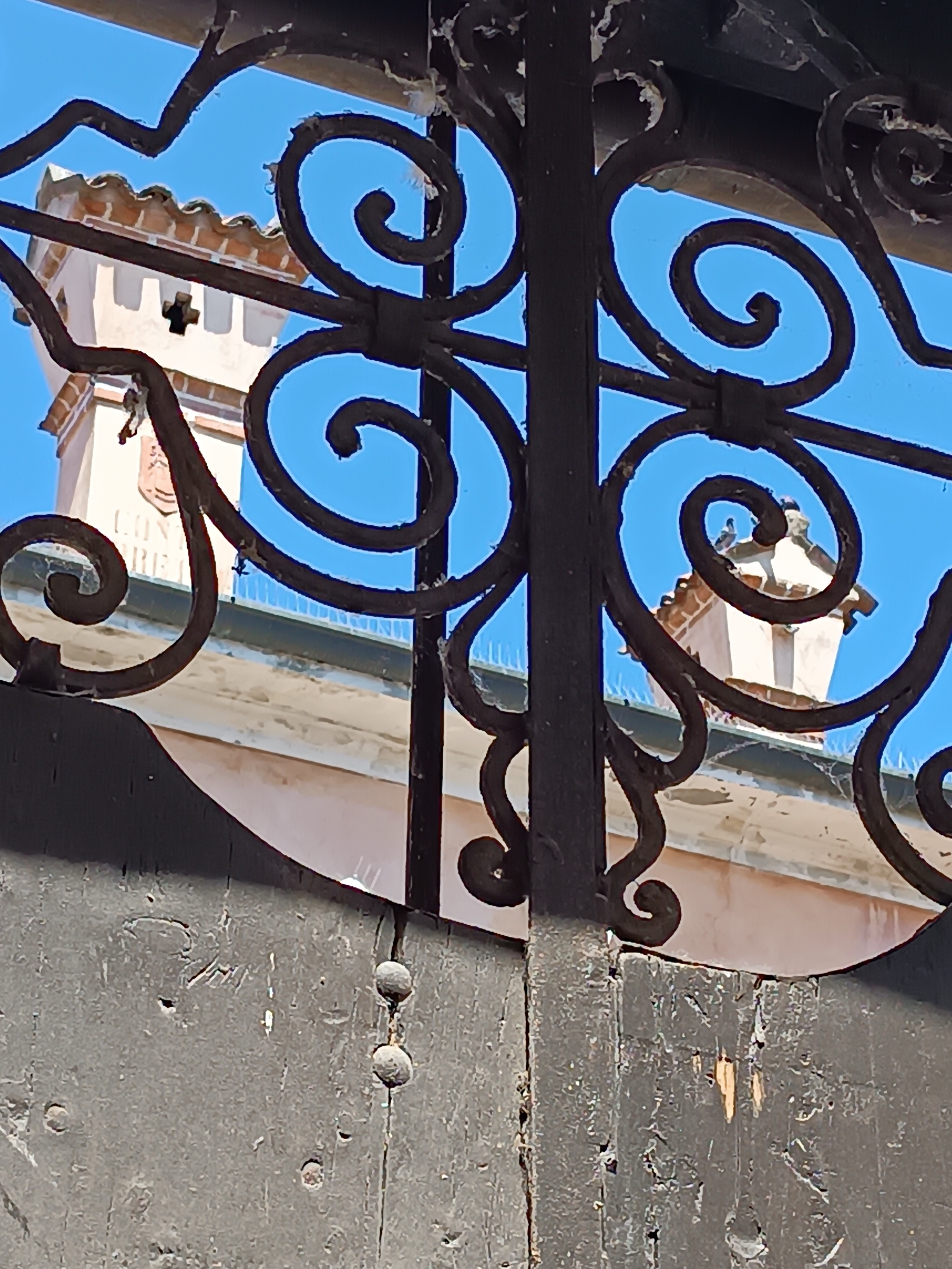 A close-up shot reveals a wrought iron gate obscuring a building in Mezzana Sant'Antonio, Italy. The gate, painted a dark color, displays ornate spiral patterns. Through the gate's openings, a light pink building with a tower is visible against a clear blue sky. The tower is adorned with a coat-of-arms or similar emblem. Bird deterrent spikes run along the building's roof. The wooden part of the gate is weathered with peeling paint.