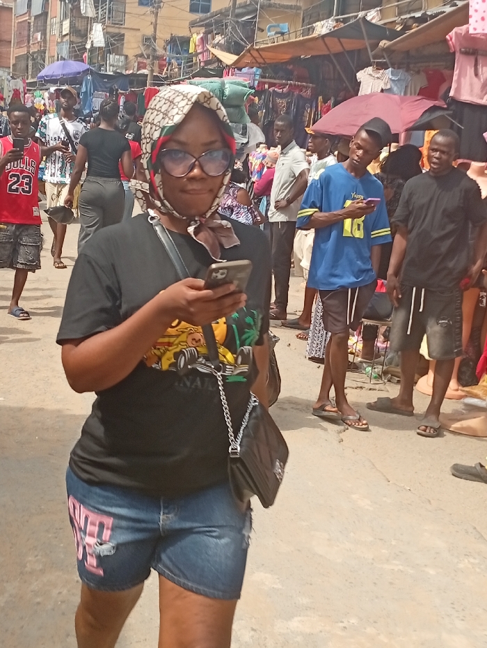 The image displays an outdoor market street scene in Fegge, Nigeria, under daylight conditions. A woman is positioned in the foreground, walking towards the viewer while looking down at a silver-colored smartphone held in both hands. She wears a cream and brown patterned headscarf with green and red accents, large dark-rimmed eyeglasses, a black short-sleeved t-shirt with a colorful graphic (featuring a yellow race car and green palm trees), and distressed blue denim shorts with pink lettering on the left leg. A black shoulder bag with a silver chain strap hangs from her left shoulder.
The background reveals a bustling urban environment. Numerous individuals, predominantly male, are present, either walking or standing. Several individuals are engaged with their mobile phones. Notable figures include a man on the left wearing a red basketball jersey with "BULLS 23," and a man in a blue t-shirt with yellow accents also looking at a phone further back. Another man in a black t-shirt stands to the right.
The street is lined with commercial activity, featuring makeshift market stalls displaying clothing and other goods under various tarpaulins and umbrellas. Multi-story buildings, some with visible balconies and clothes hanging, form the backdrop. The ground surface appears to be light-colored concrete or paved earth.