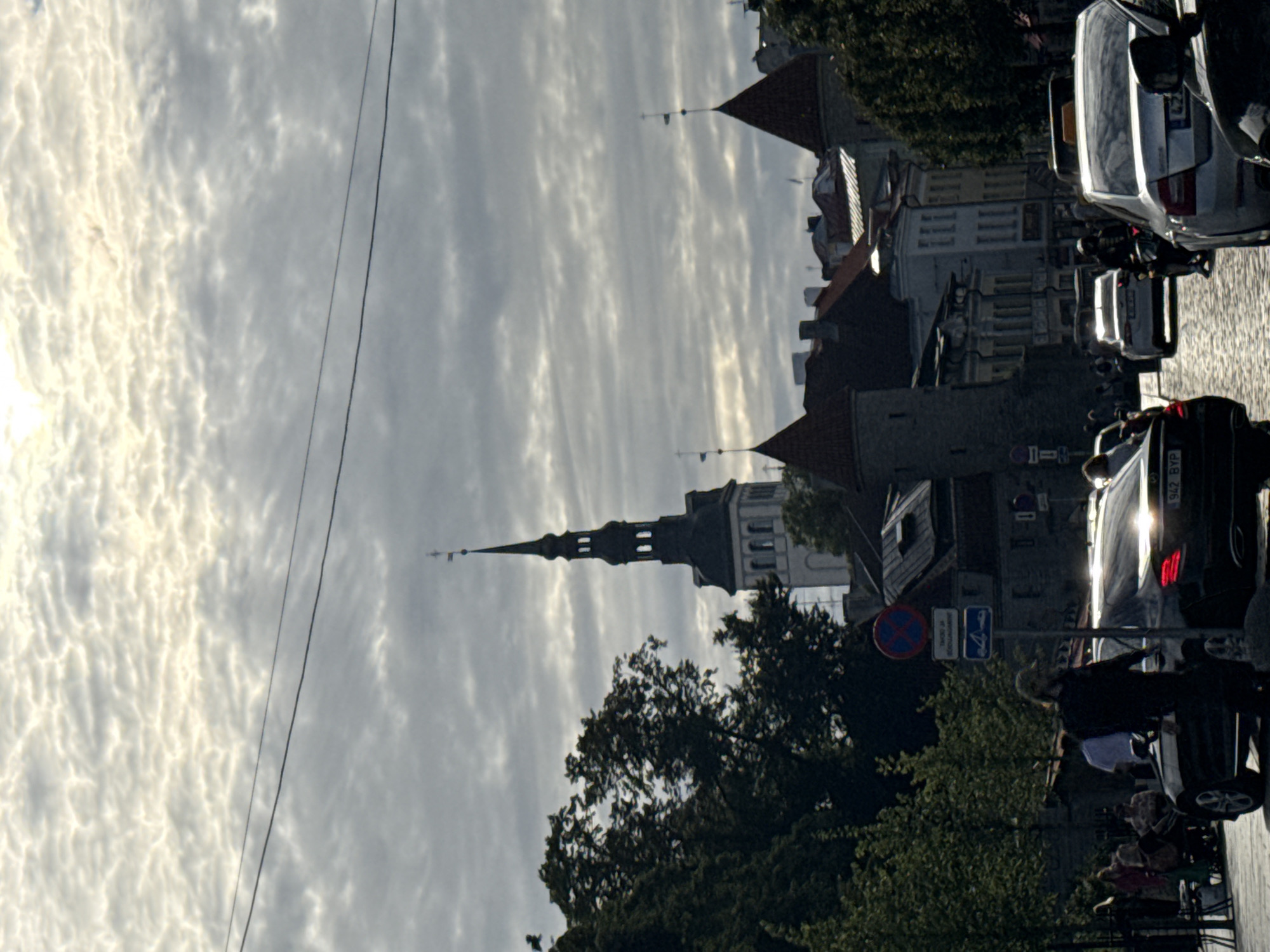 The original image is a vertical, medium-shot photograph taken in an urban environment, likely a city street, featuring a street scene with architectural elements and vehicles. The sky is overcast with dramatic, textured clouds that are illuminated by the sun, creating a bright, diffused light. In the background, a tall, dark-spired church tower dominates the skyline, with other buildings featuring conical roofs visible on the right side of the frame.
In the mid-ground, a street is lined with buildings and trees. Several cars are present on the street, including a black sedan in the foreground with its brake lights illuminated, and other vehicles further down the road, some appearing to be taxis due to yellow roof signs. A motorcycle is visible between two cars. People are scattered throughout the scene, some standing on the sidewalk and others near the cars. One person, dressed in dark clothing and wearing a cap, is standing next to the black sedan in the foreground, seemingly observing. Another person, dressed in a light-colored shirt, is also visible near the cars. A traffic signpost with blue and red circular signs is positioned on the left side of the street.
The foreground features a cobblestone street. The overall impression is a busy street scene in a European city, possibly in the late afternoon or early evening, given the quality of light and the presence of illuminated vehicle lights. The image has a slightly grainy texture, suggesting it may have been taken with a mobile device or in low-light conditions.