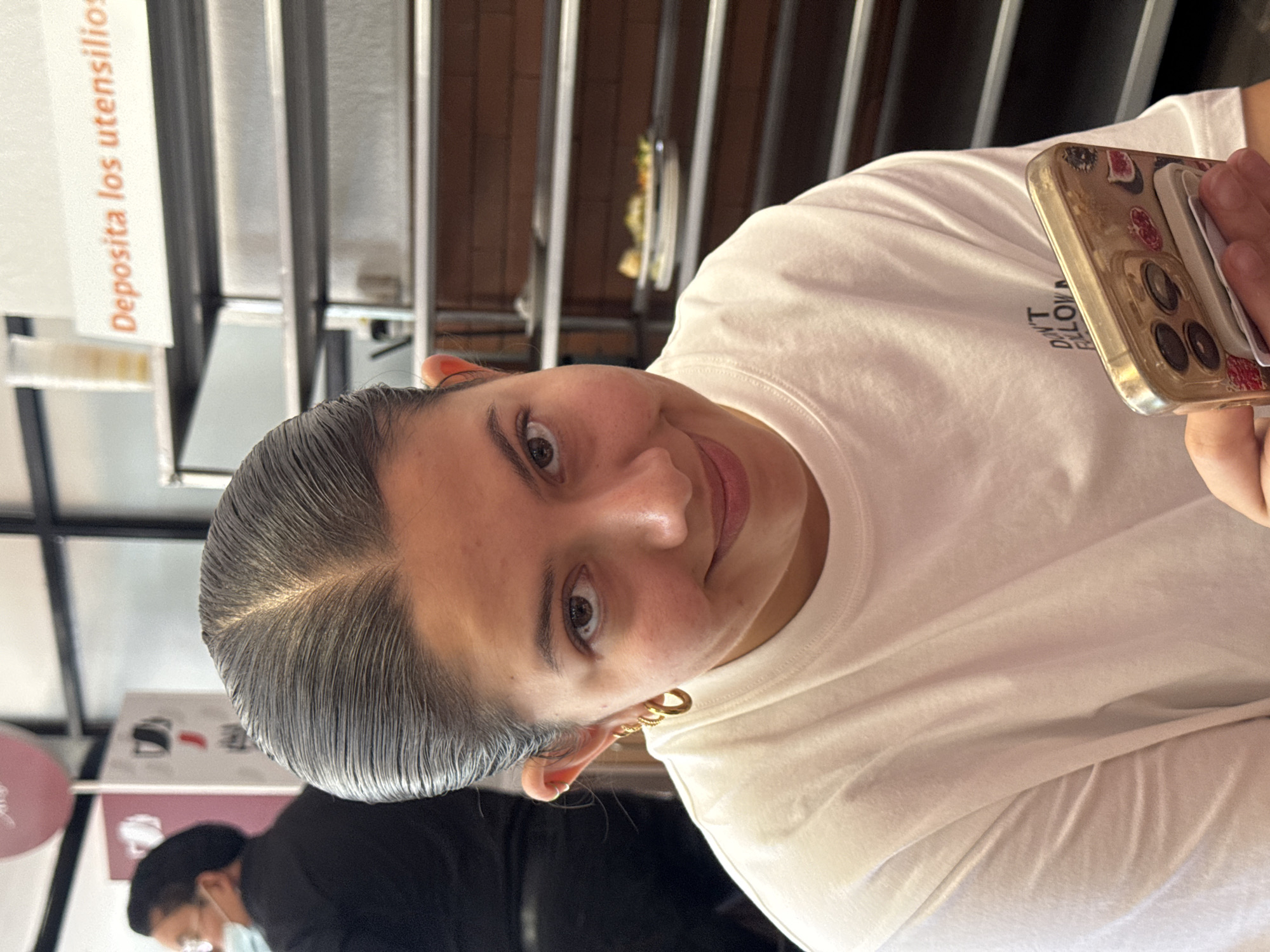 A woman with dark hair pulled back and wearing gold hoop earrings is looking at the camera and smiling. She is wearing a white t-shirt. She is holding a gold-colored smartphone with a fruit-themed case in her right hand. The phone screen is not visible. A person wearing a black shirt and a face mask is partially visible in the background. The background also includes a metal structure with a sign in Spanish, "Deposita los utensilios". The scene appears to be indoors, possibly a restaurant or food establishment. The location is San Andrés Cholula, Mexico.