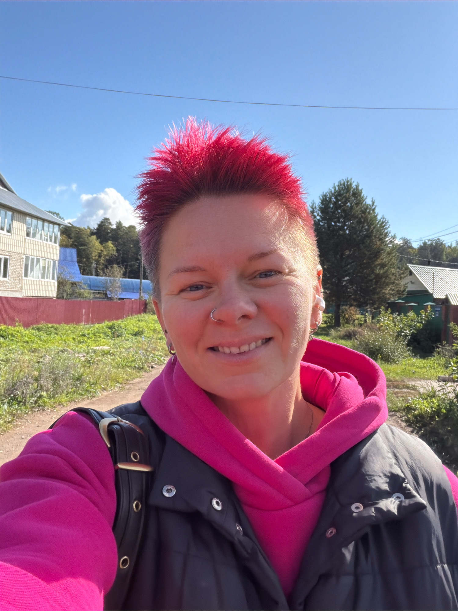 A woman with bright pink spiky hair, a nose ring, and earrings smiles at the camera. She is wearing a pink hoodie and a black vest, and a black bag is slung over her shoulder. The background shows a dirt road, a red fence, green bushes and trees, and buildings in the distance under a clear blue sky. This scene appears to be on a sunny day in the city of Chaikovsky, Russia.
