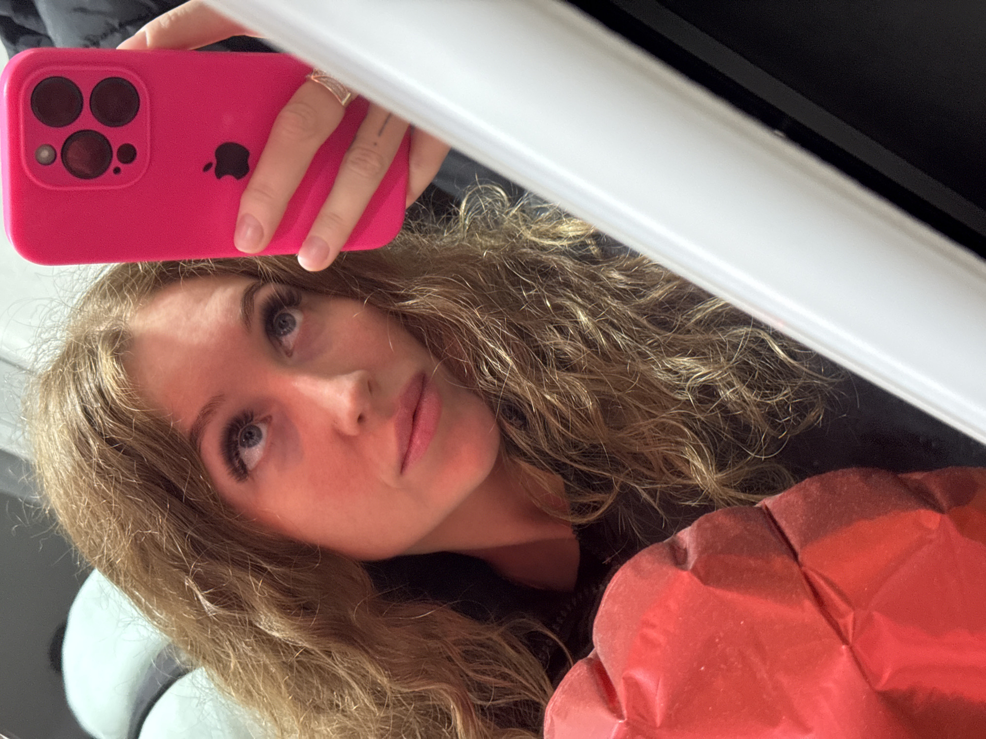 A young woman with blonde curly hair is taking a selfie using a pink iPhone in what appears to be an indoor environment. She is looking at the camera with a slight upward angle. The background includes a white object, possibly a frame or door, and some undefined elements behind her. The red puffy material in the bottom right might be a jacket. There is no information in the image to determine the location in Chaikovsky, Russia.